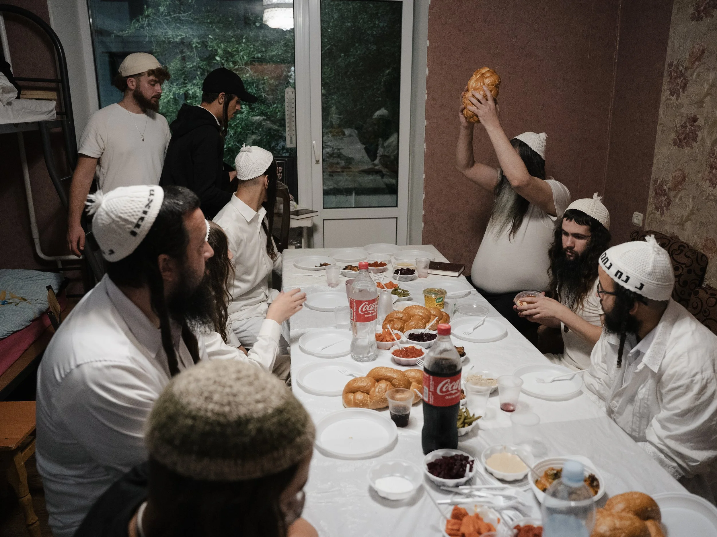 A group of people gathered around a table for a meal, with some wearing white knitted caps. One person is holding a large loaf of bread, raised above their head. The table has various dishes including bread, salad, and drinks.