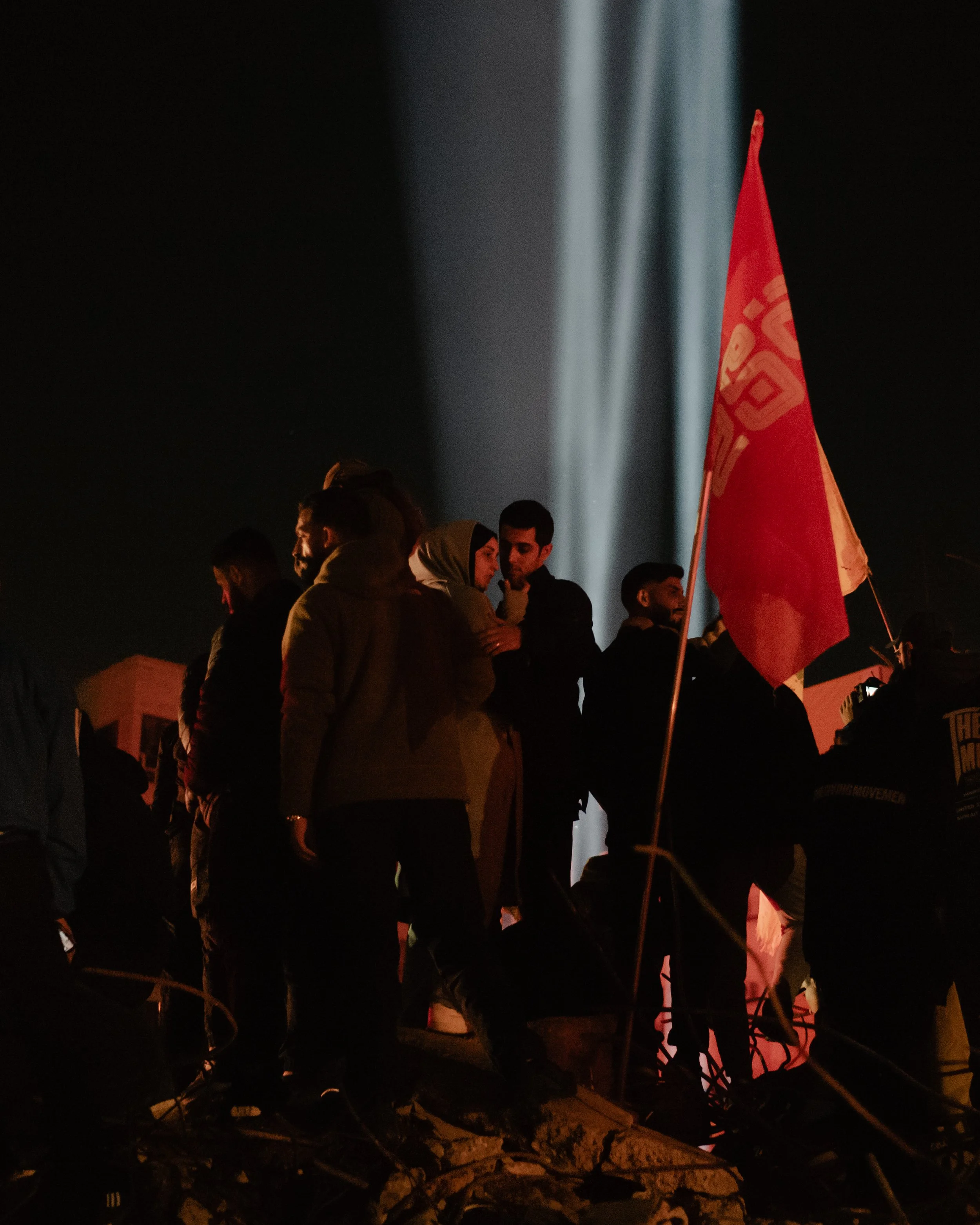 People gathered at night holding a red flag with gold lettering; the setting appears to be a protest or demonstration, with shadows and a dark background.