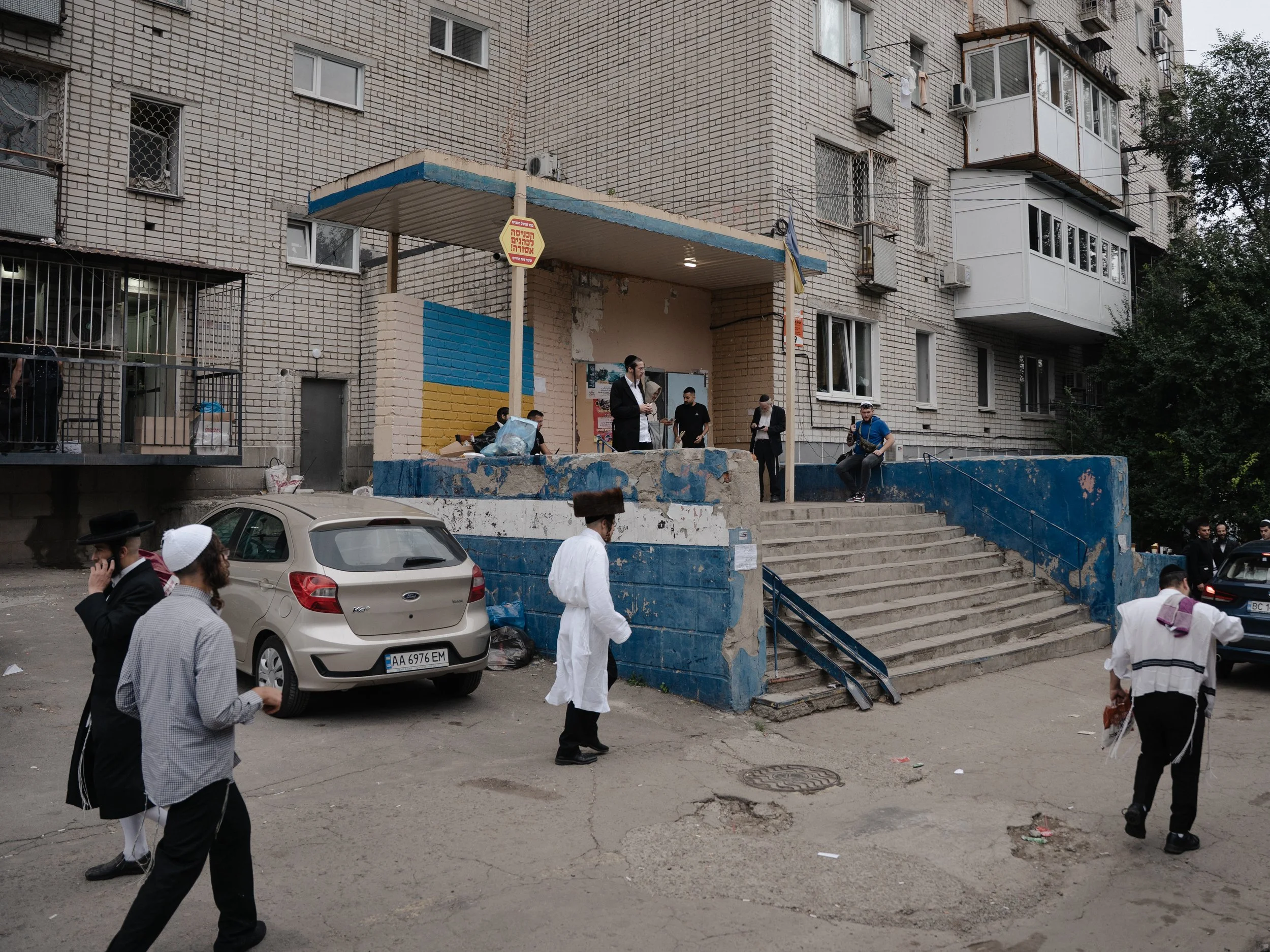People outside a multi-story brick building with a blue and yellow mural on the lower wall. Some individuals are walking, and others are gathered around the entrance. Cars are parked in front of the building.
