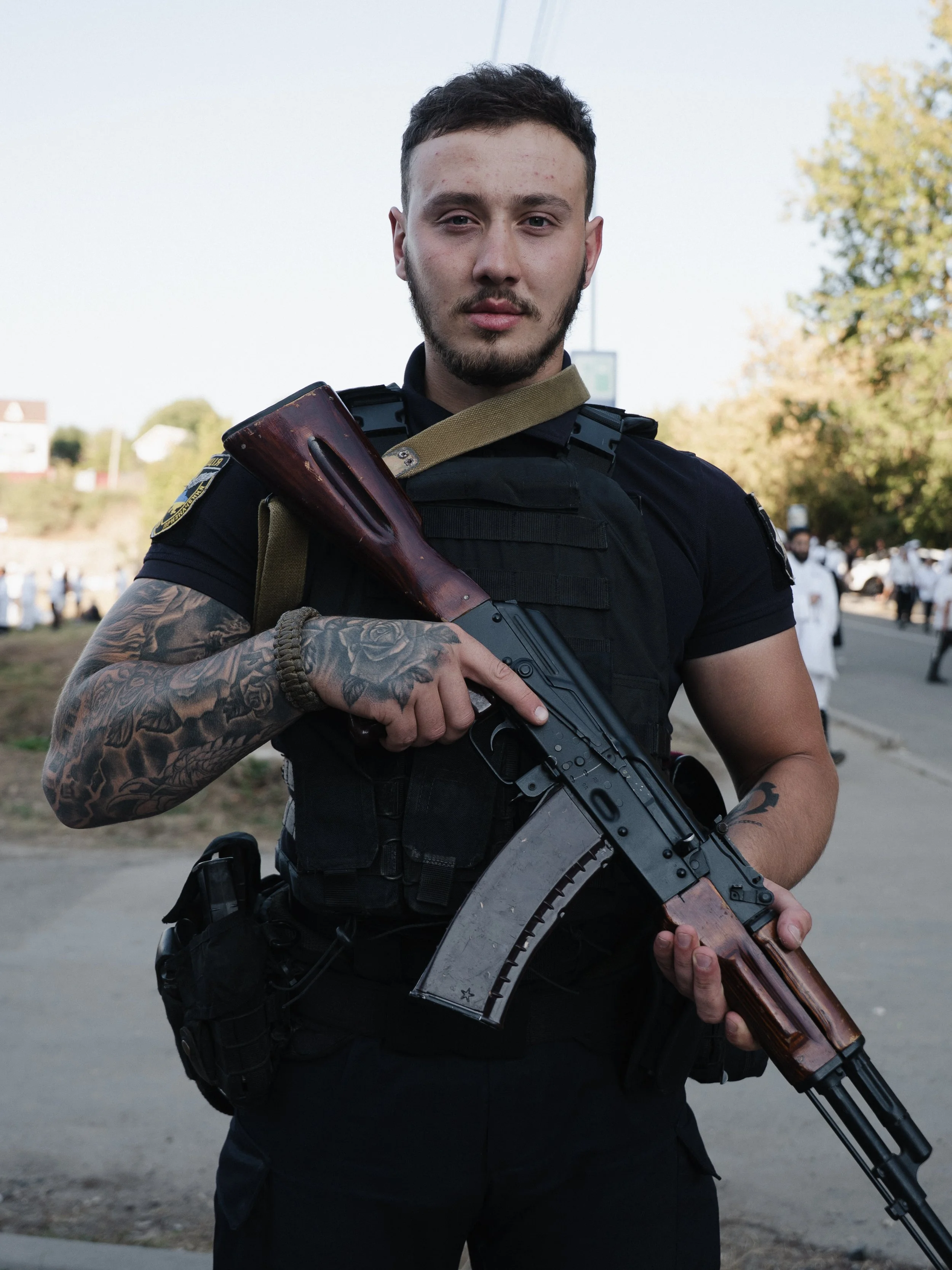 A police officer holding a rifle outside on a street with trees and people in the background.