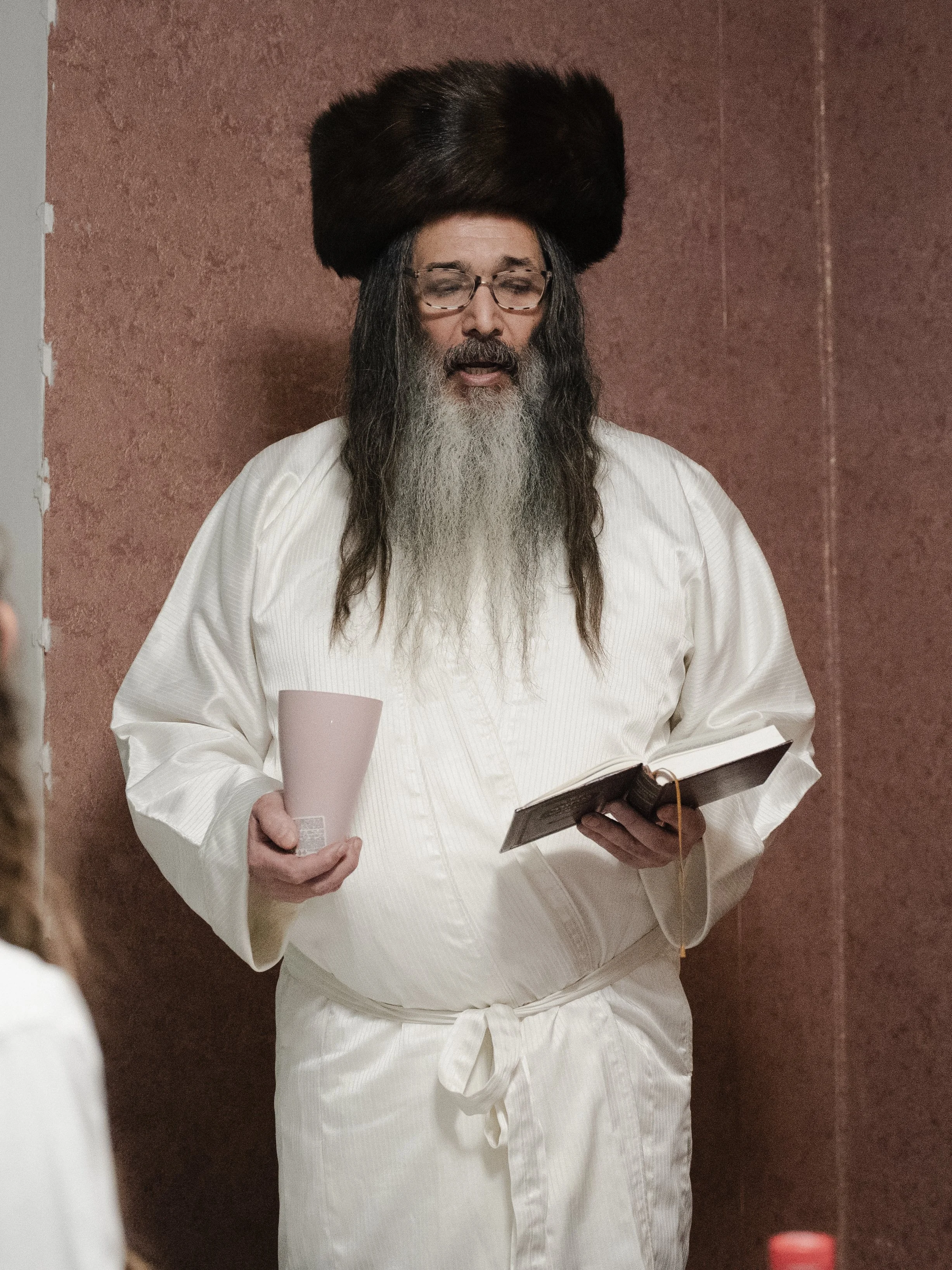 A man with long hair, a beard, and glasses wearing traditional Jewish clothing, including a large fur hat called a shtreimel, holding a pink cup and reading from a book in a room with reddish walls.
