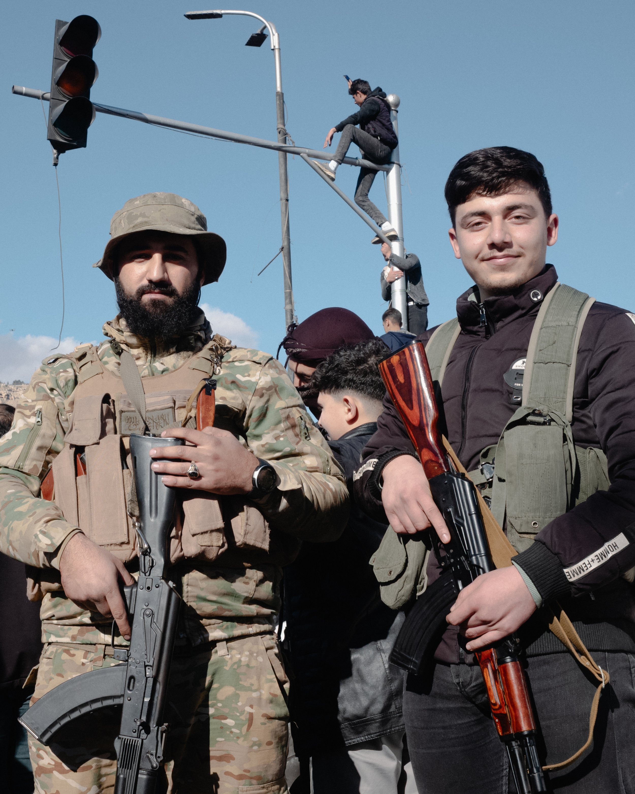 Two young men standing side by side holding rifles, with a background of a traffic light and a person sitting on a utility pole, against a clear blue sky.