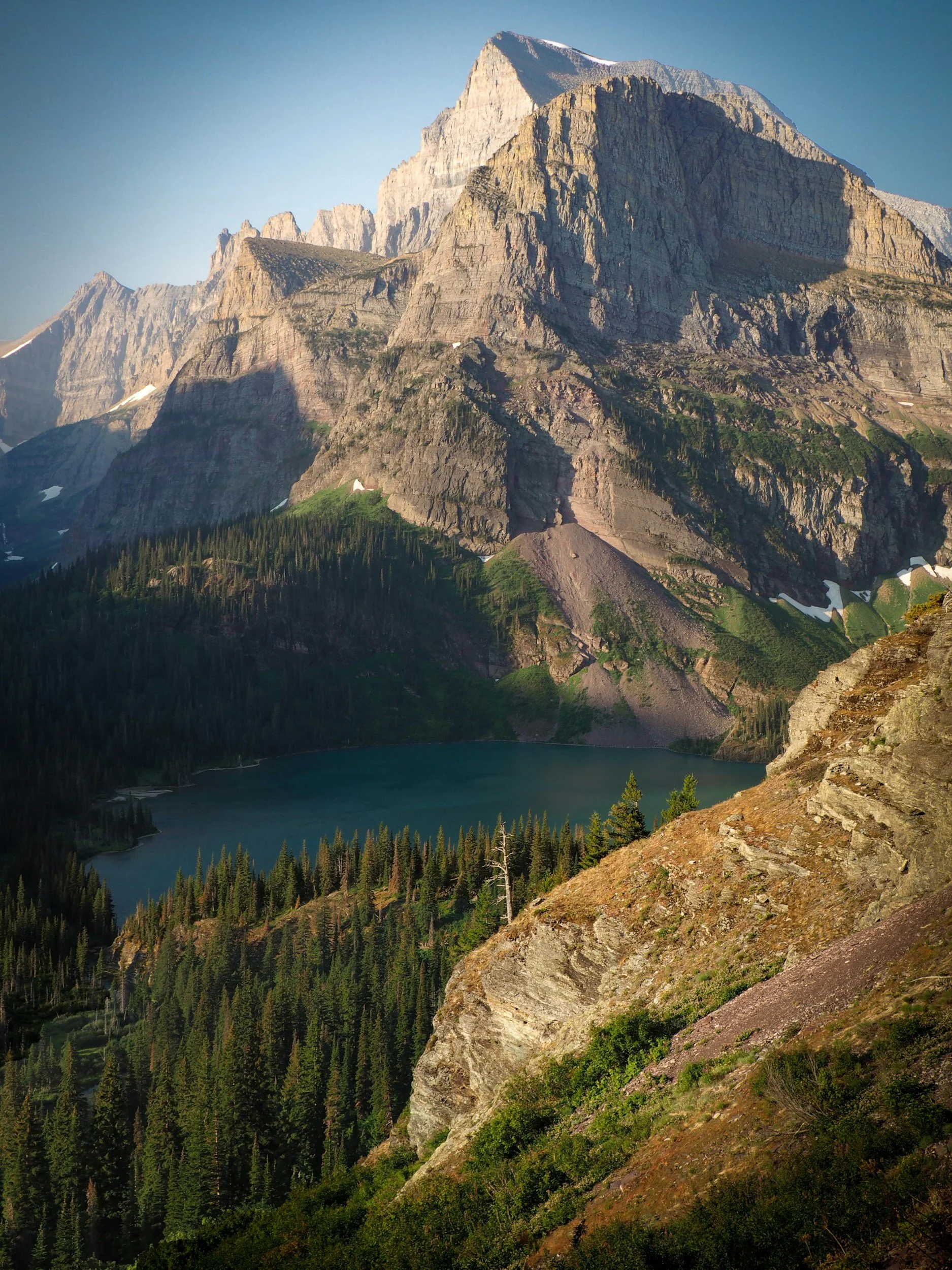 A large mountain with rocky cliffs and a snow-capped peak, surrounded by green trees and a lake at the base.