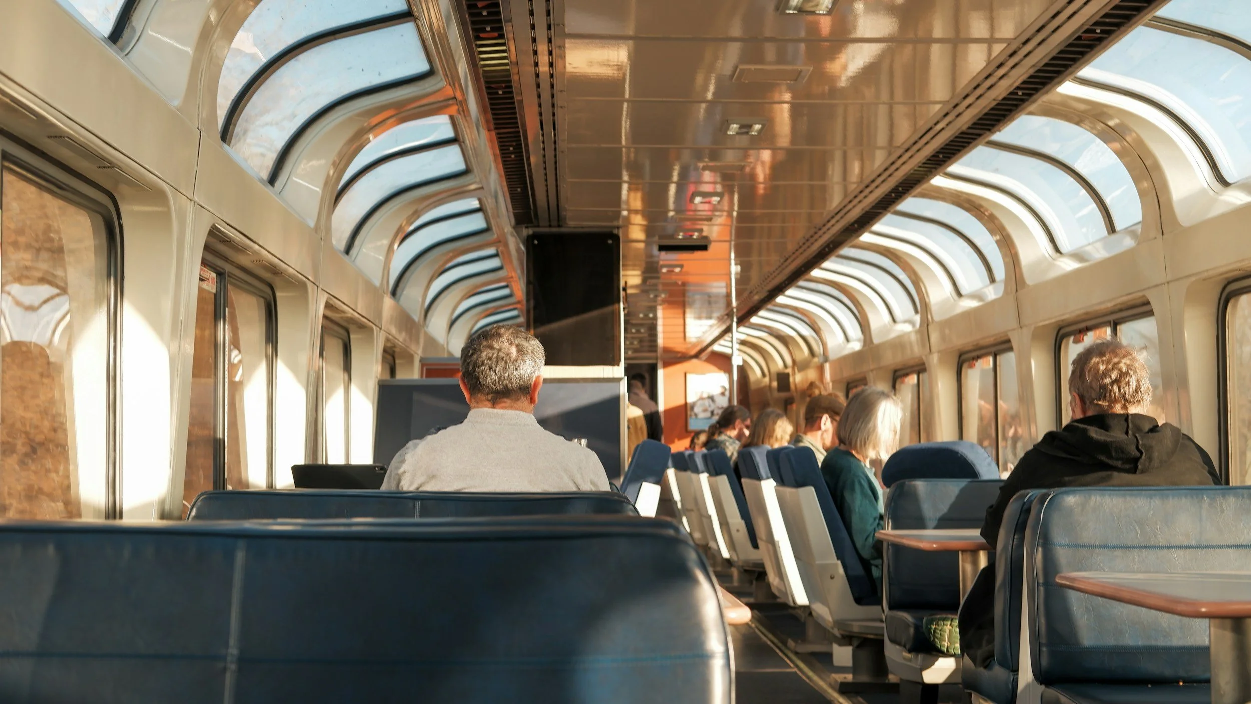 Passengers sitting inside a train with large windows and arched glass roof, during daytime.