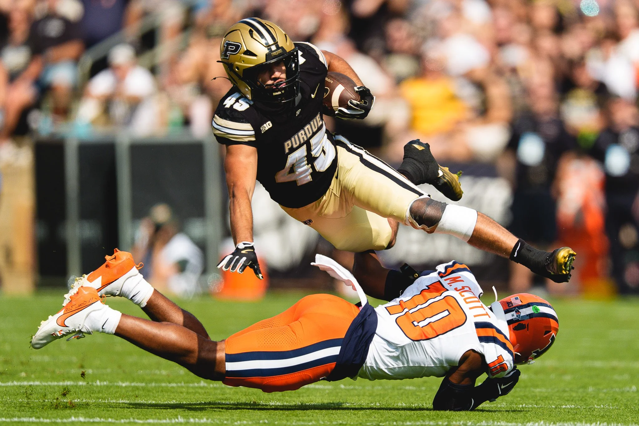 An American football game showing a Purdue player in black and gold uniforms jumping over a player in an orange and navy uniform who is on the ground trying to tackle him.