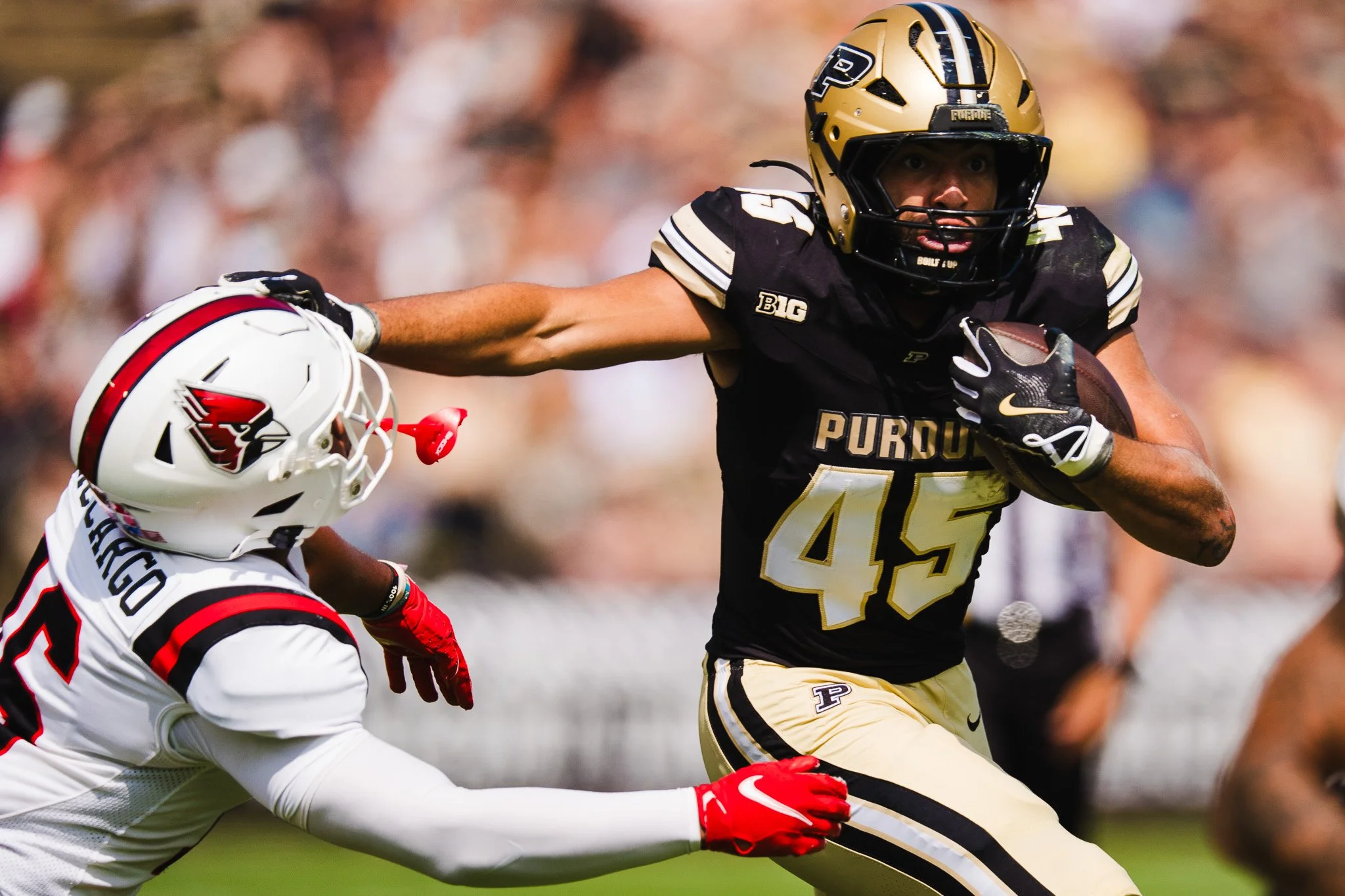 A college football player from Purdue University running with the ball while being tackled by a player from the opposing team wearing a white helmet and jersey.