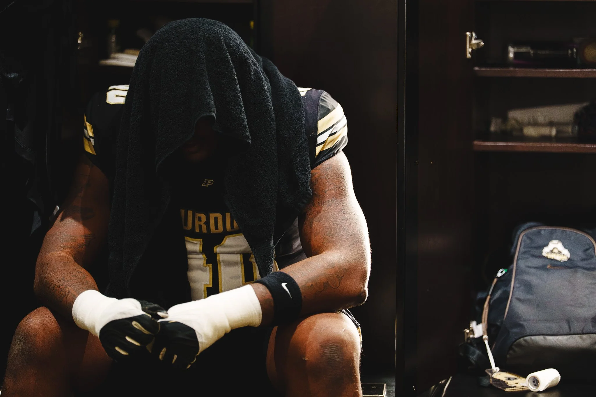 A football player with a towel over his head, sitting in a locker room, wearing a black and yellow uniform with the word 'PURDUE' visible on the jersey, gloves, and wristbands, with a backpack and locker nearby.