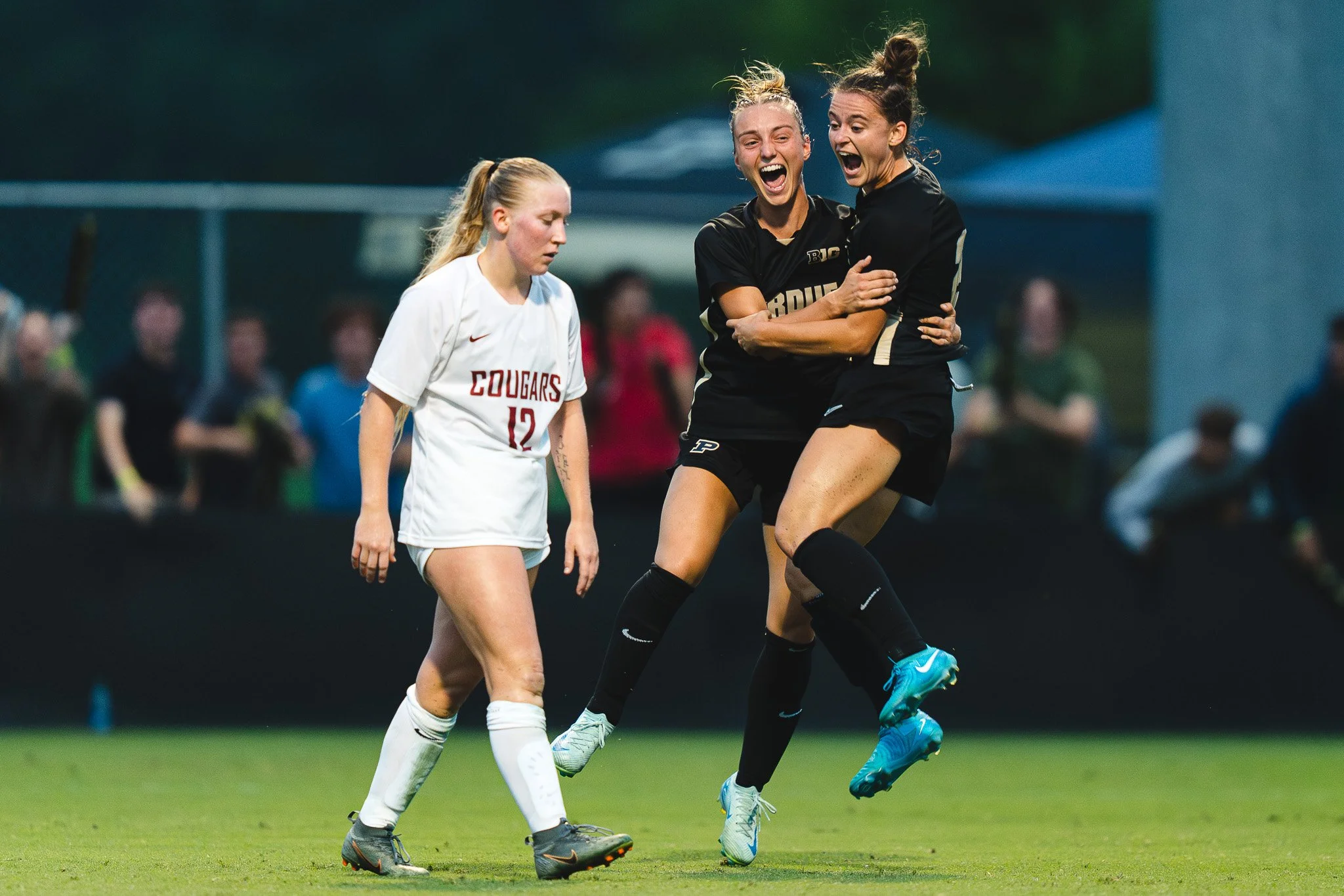 Two female soccer players in black uniforms are celebrating and hugging each other on the field. A third female player in a white uniform with the number 12 and 