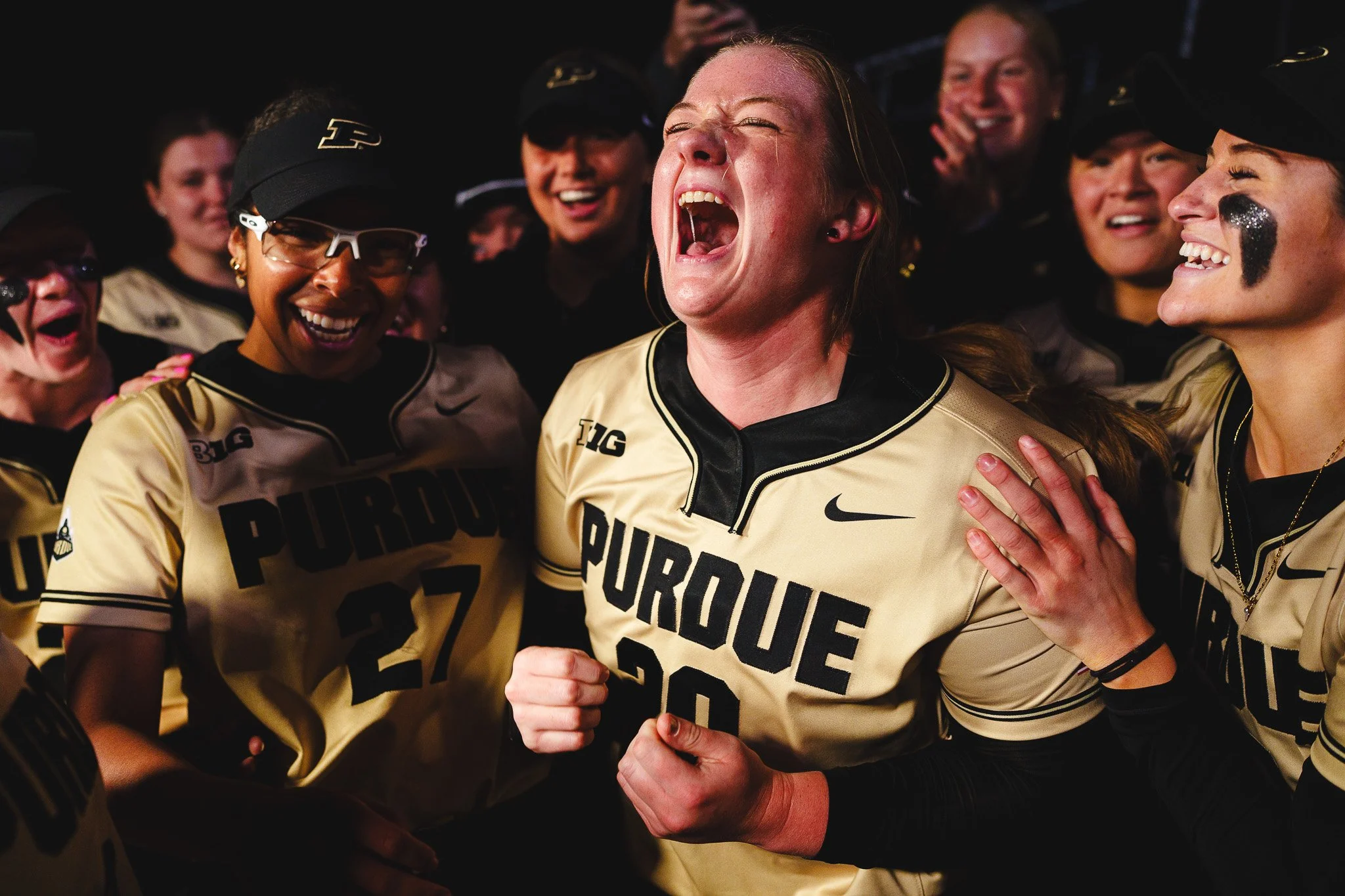 Women in Purdue football jerseys celebrating together, with one woman in the center shouting with joy and others smiling around her.