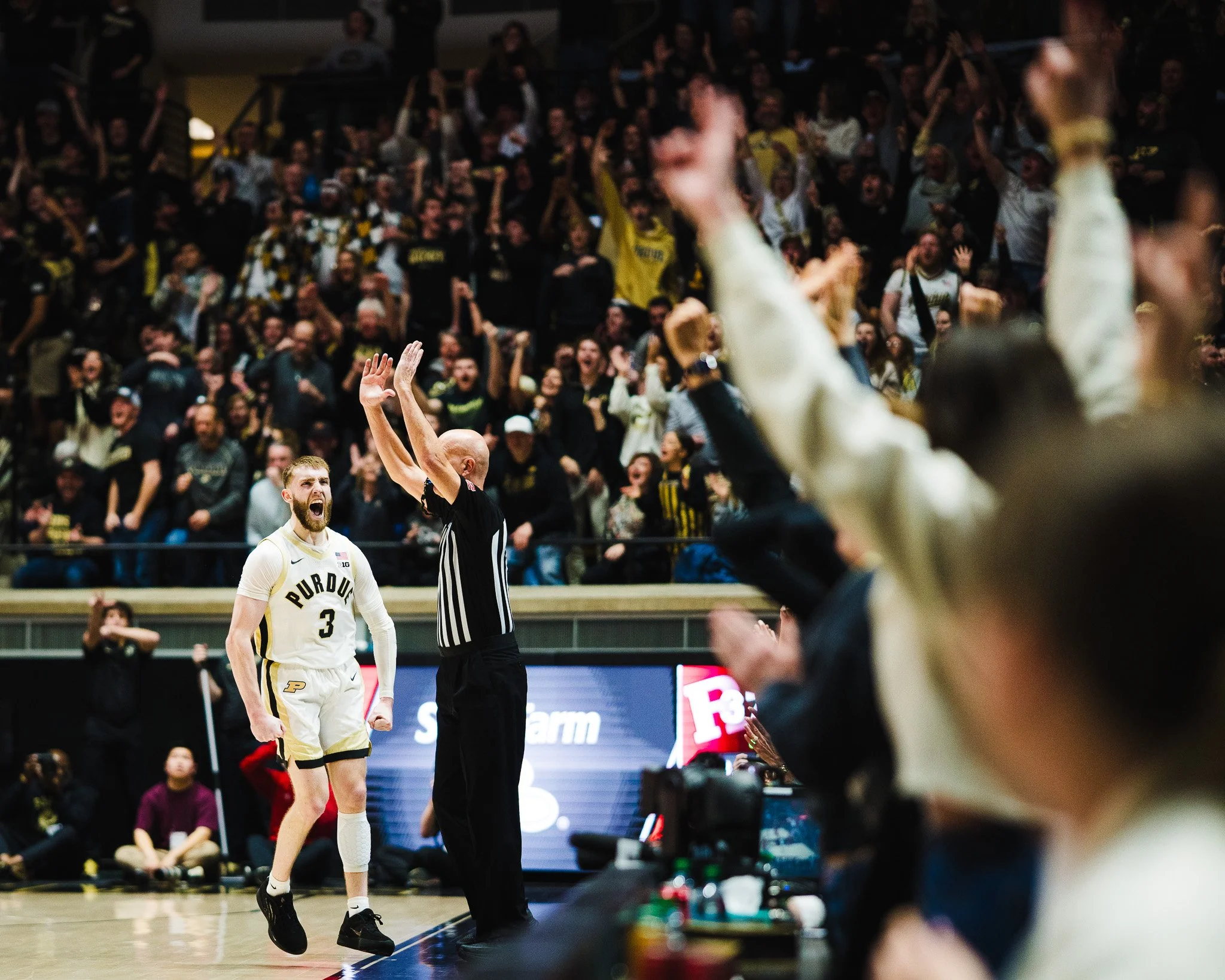 Purdue basketball player with number 3 celebrating after a successful play as the referee raises his hand, with cheering spectators in the background.