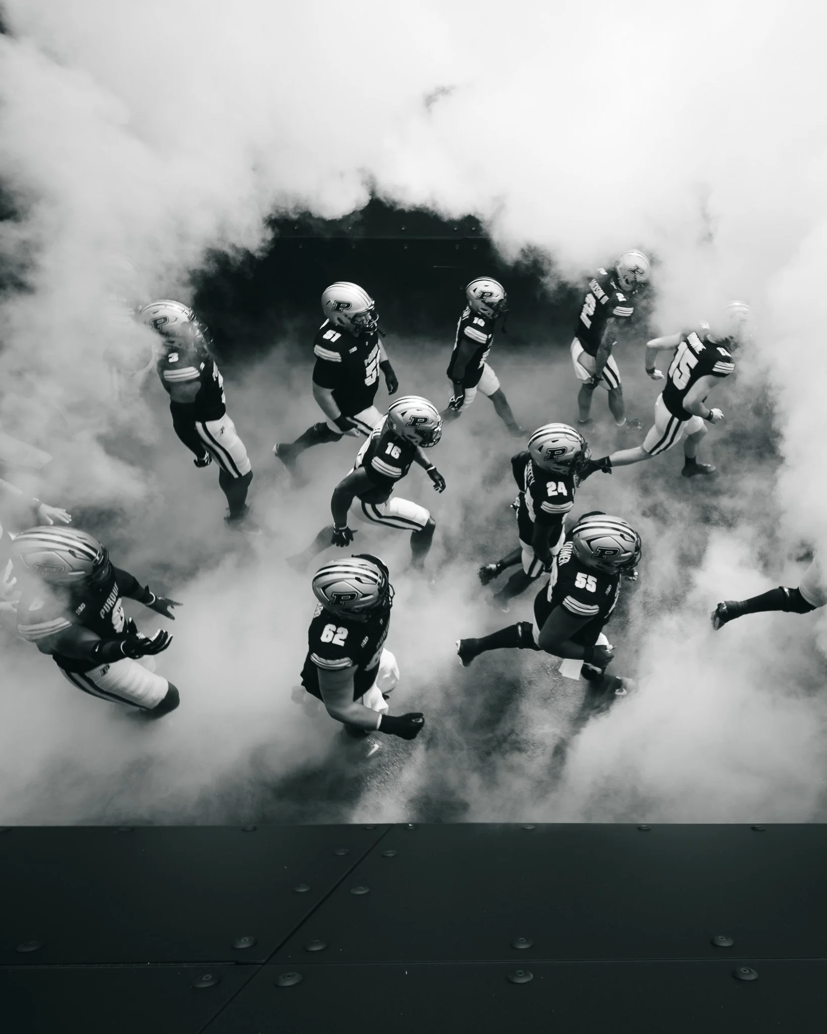 Black and white photo of football players running through a smoky tunnel holding hands.