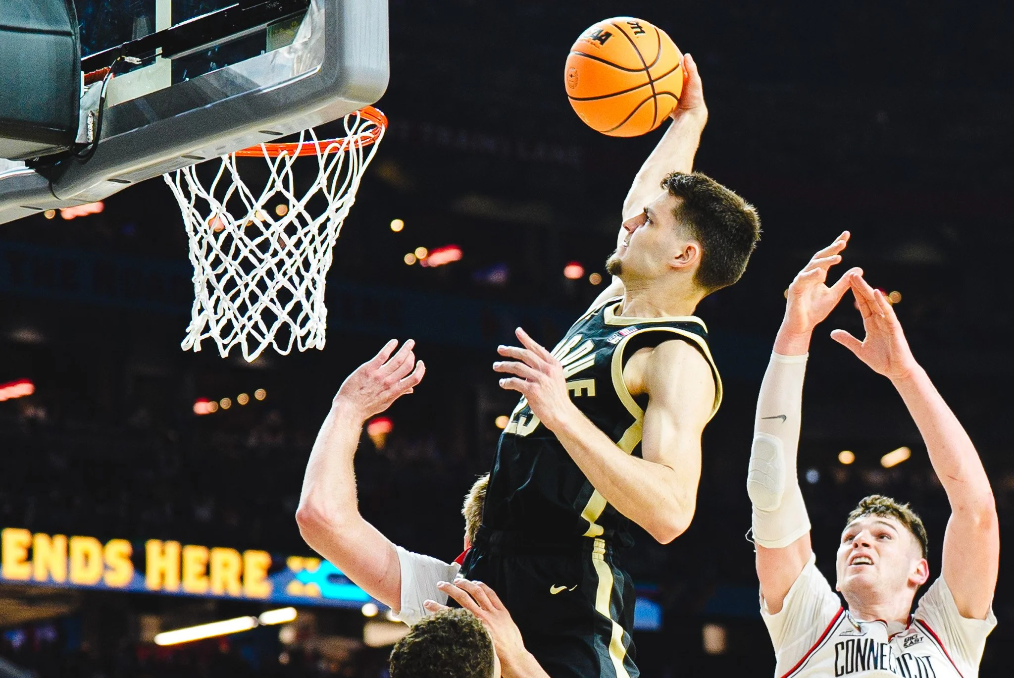 A basketball player in a black and gold uniform is jumping towards the basket for a shot, with another player in a white jersey attempting to block him during a game.