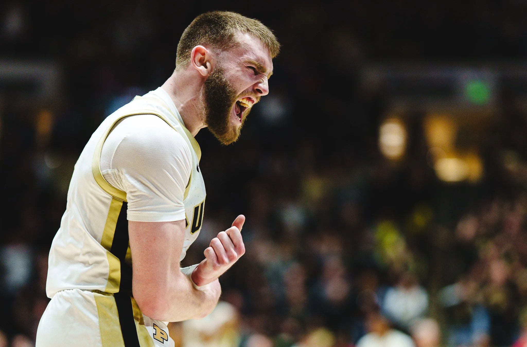 A male basketball player with a beard and short hair, wearing a white and gold jersey, celebrating on the court with a joyful expression and clenched fist.