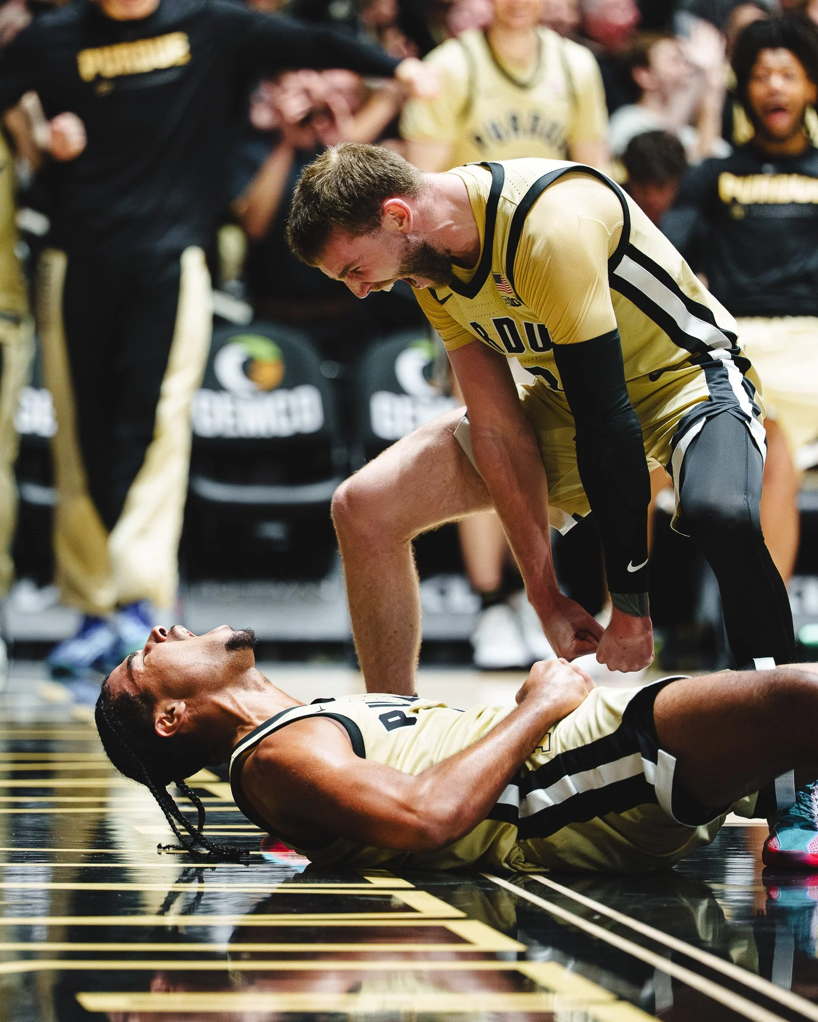 A basketball player lying on the court celebrates while another player leans over him, both wearing yellow and black uniforms, with teammates and coaches in the background.