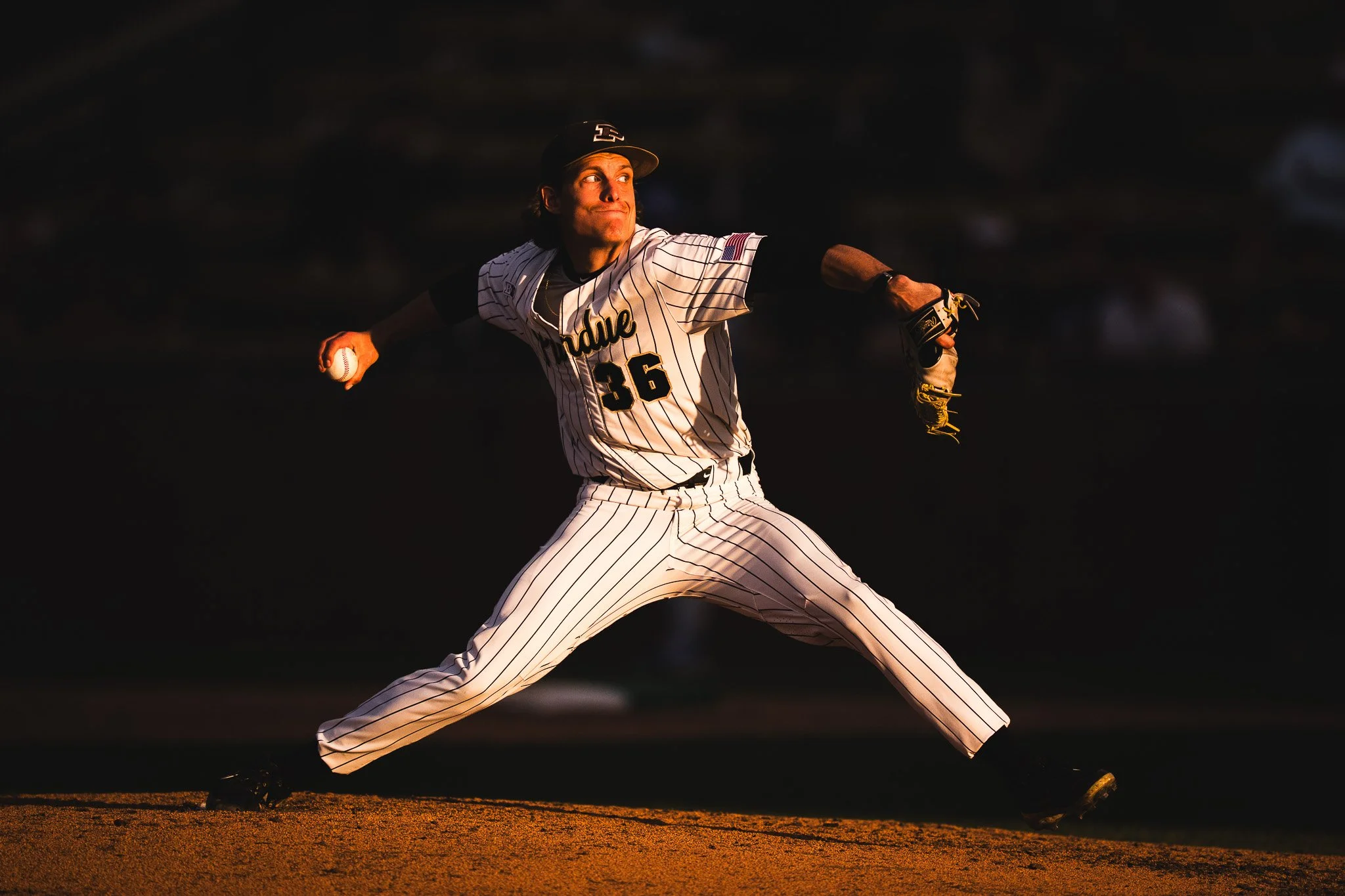 A baseball pitcher in a white and black striped uniform tossing a ball on the field during sunset.