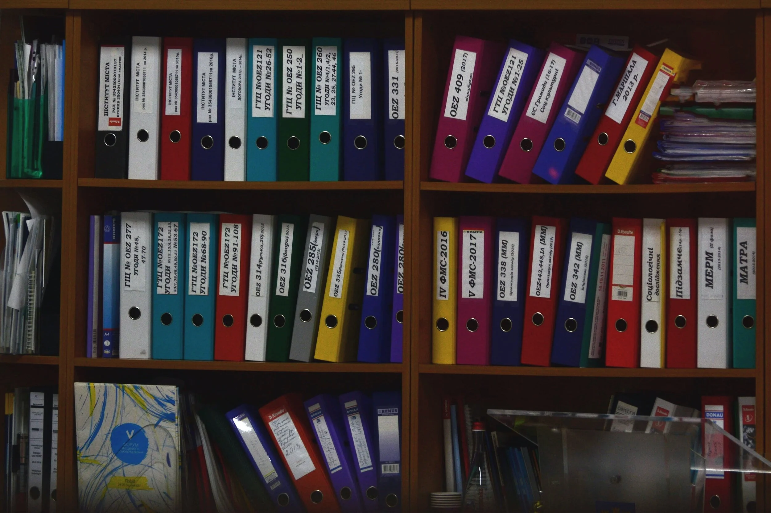 A bookshelf filled with colorful binders organized in rows, each labeled with text, some with Cyrillic characters. Documents and folders are also visible on the shelves, adding to the organized office setting.