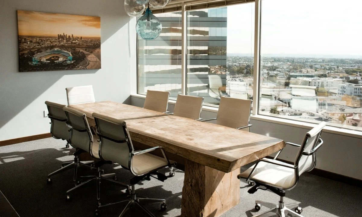 Conference room with wooden table, white office chairs, large windows, and a cityscape painting on the wall.