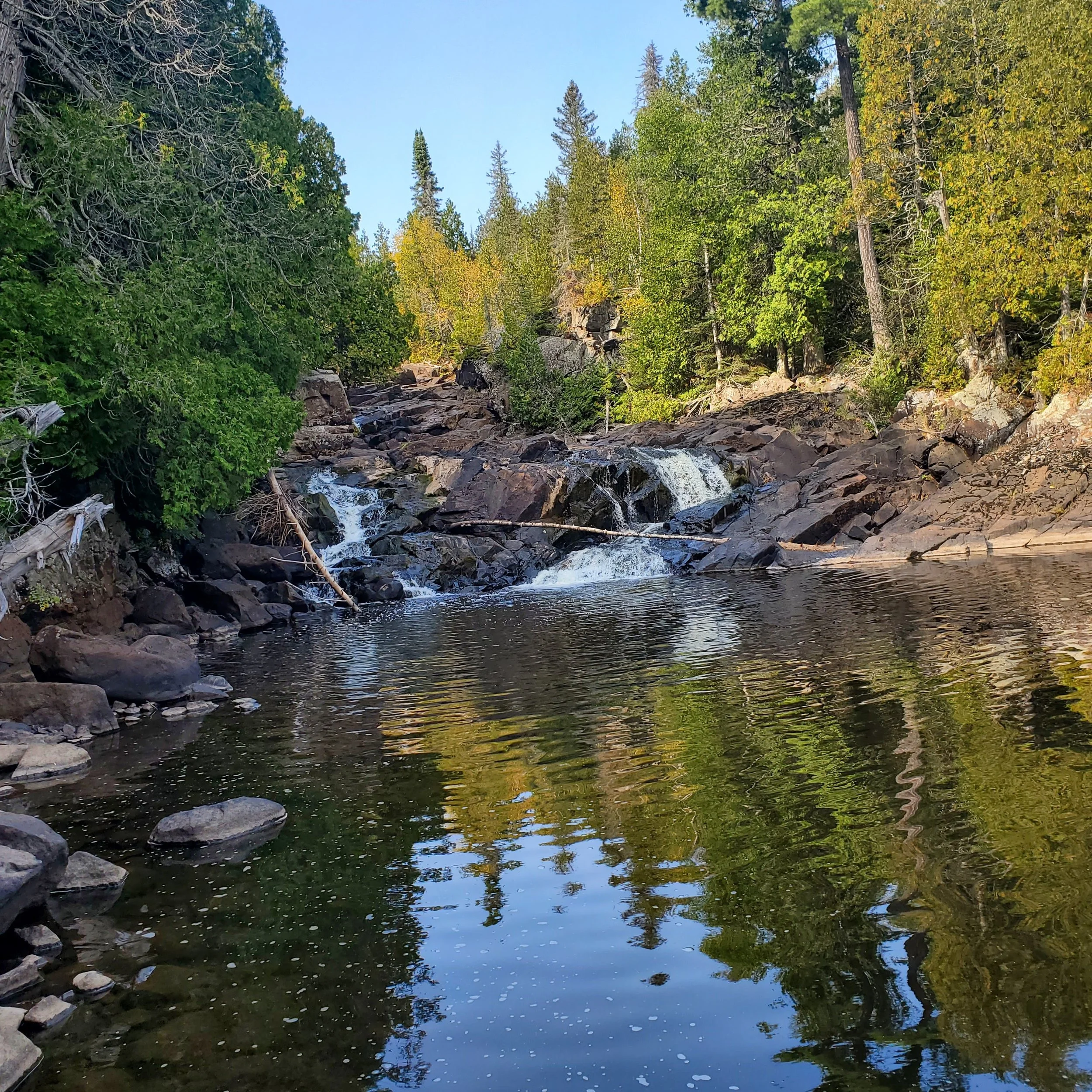 A peaceful stream flowing over rocks surrounded by green trees, under a clear blue sky at George H. Crosby Manitou state park.