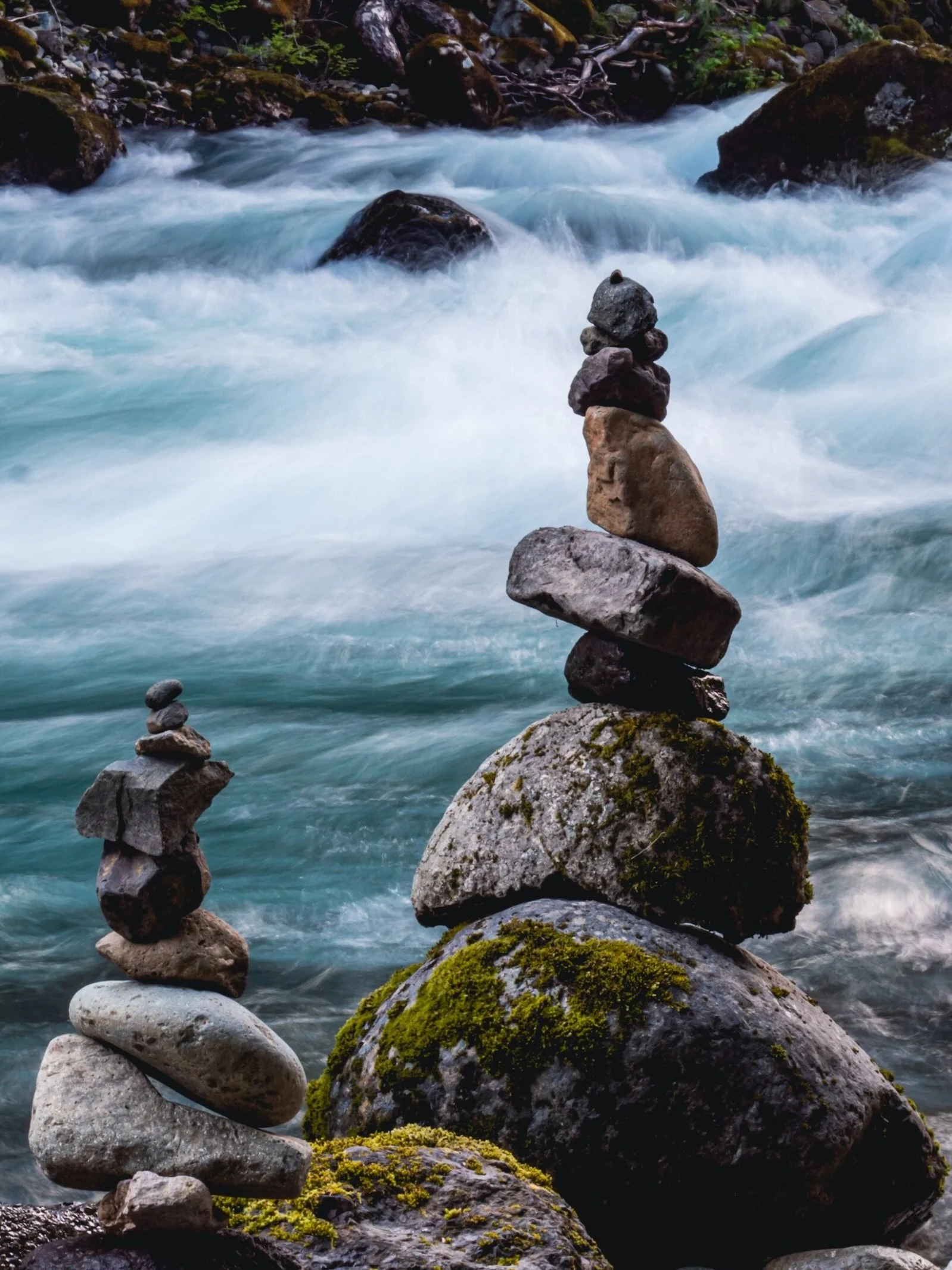 A series of balanced rocks stacked above each other at the edge of a flowing river with rocks and green moss, surrounded by water and more rocks.