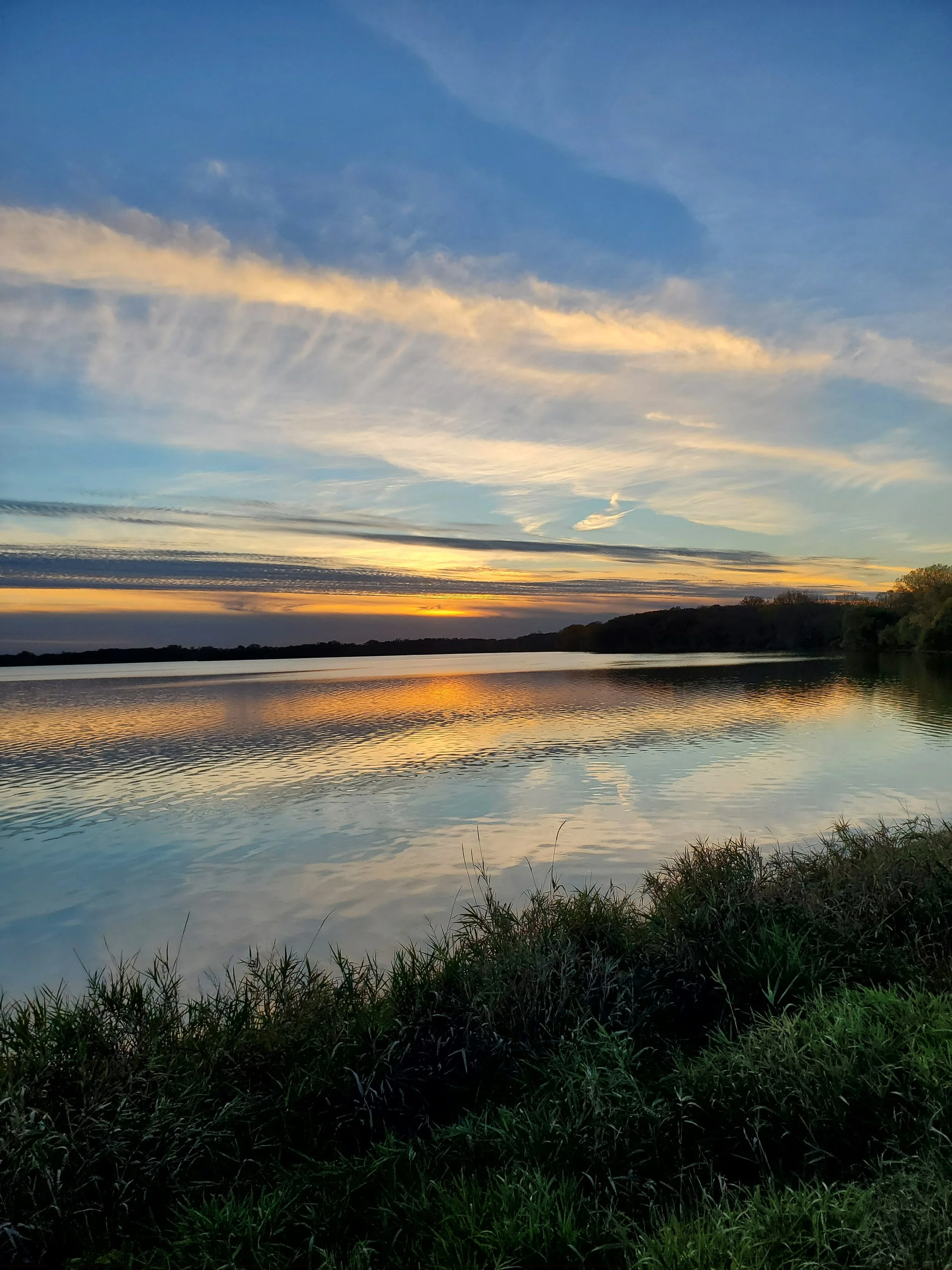 Sunset over a body of water with a sky filled with clouds, reflected in the water, with grassy shoreline in the foreground. Photo taken at Myre-Big Island state park.