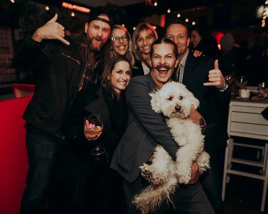 A group of friends celebrating together, one holding a small white dog, at an evening party