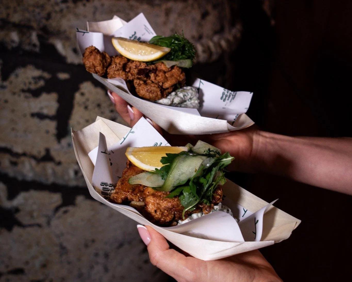 Two servings of fried chicken with lemon, cucumber and herbs in branded paper trays, a walk-and-fork option