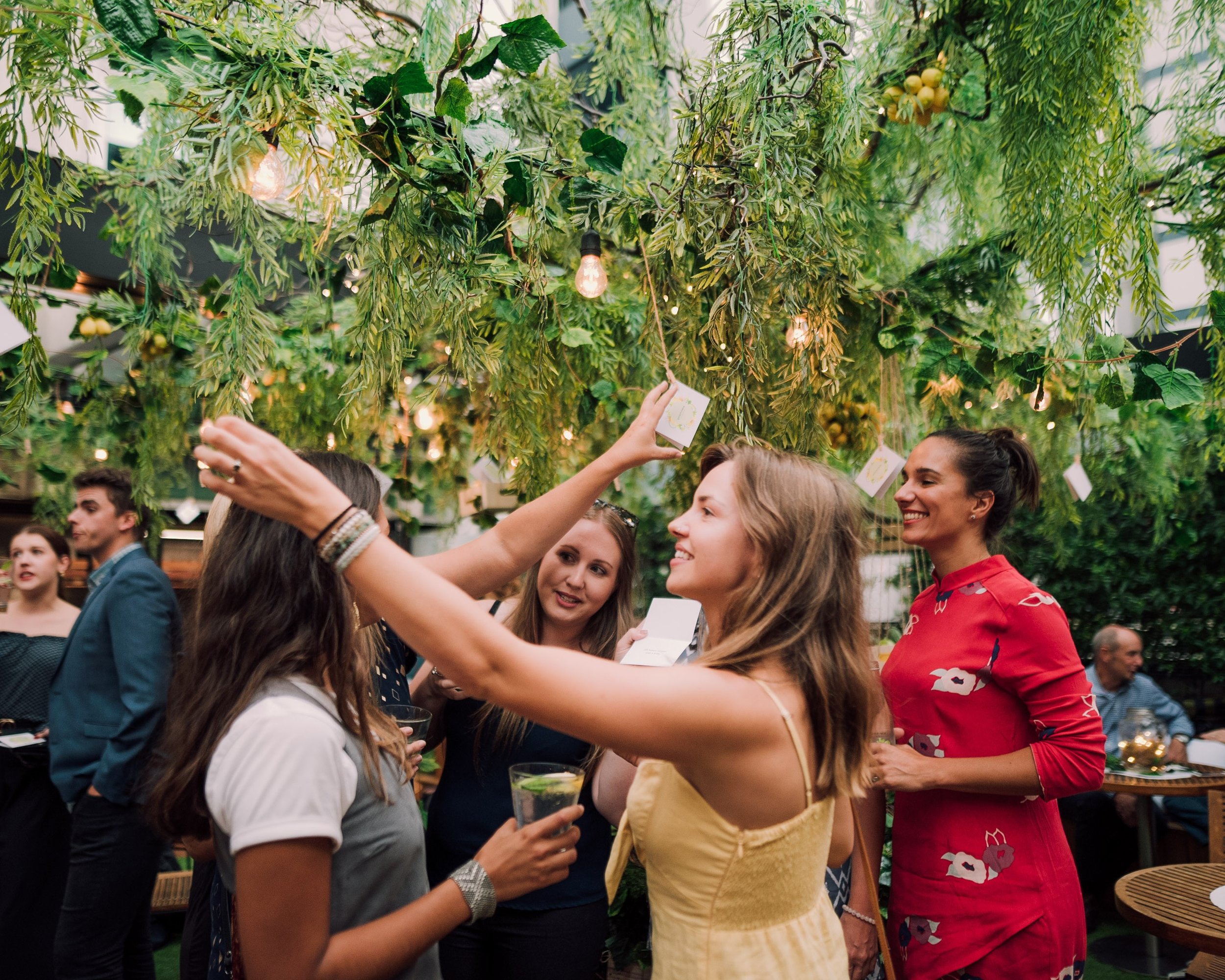 Guests at a private event reaching for cards hung from a lush greenery installation