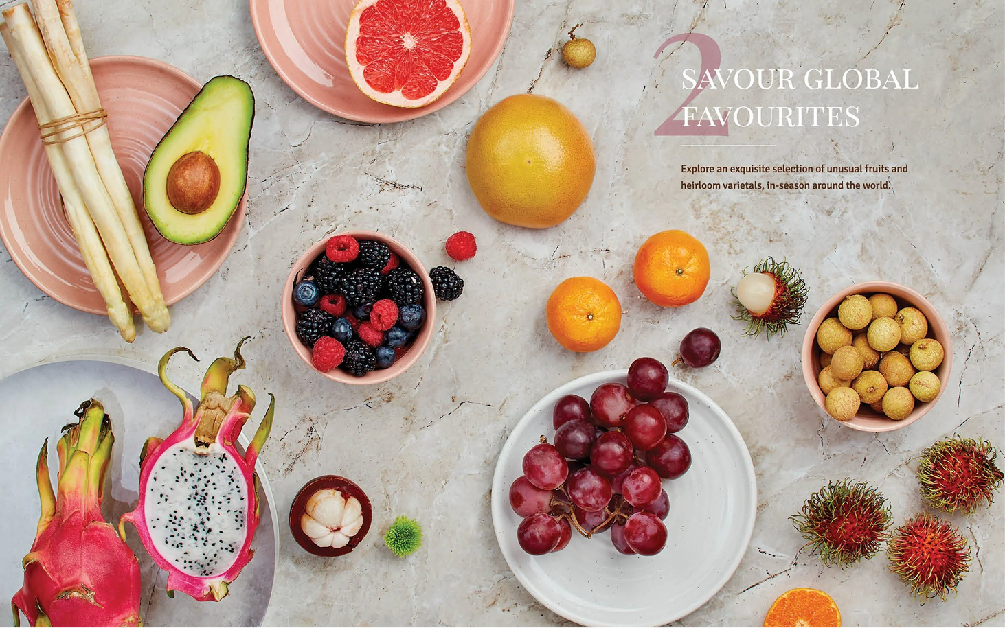 An assortment of exotic fruits on a marble surface including avocado, dragon fruit, grapes, berries, citrus, lychee, rambutan, and longan, with some plates and bowls for presentation.