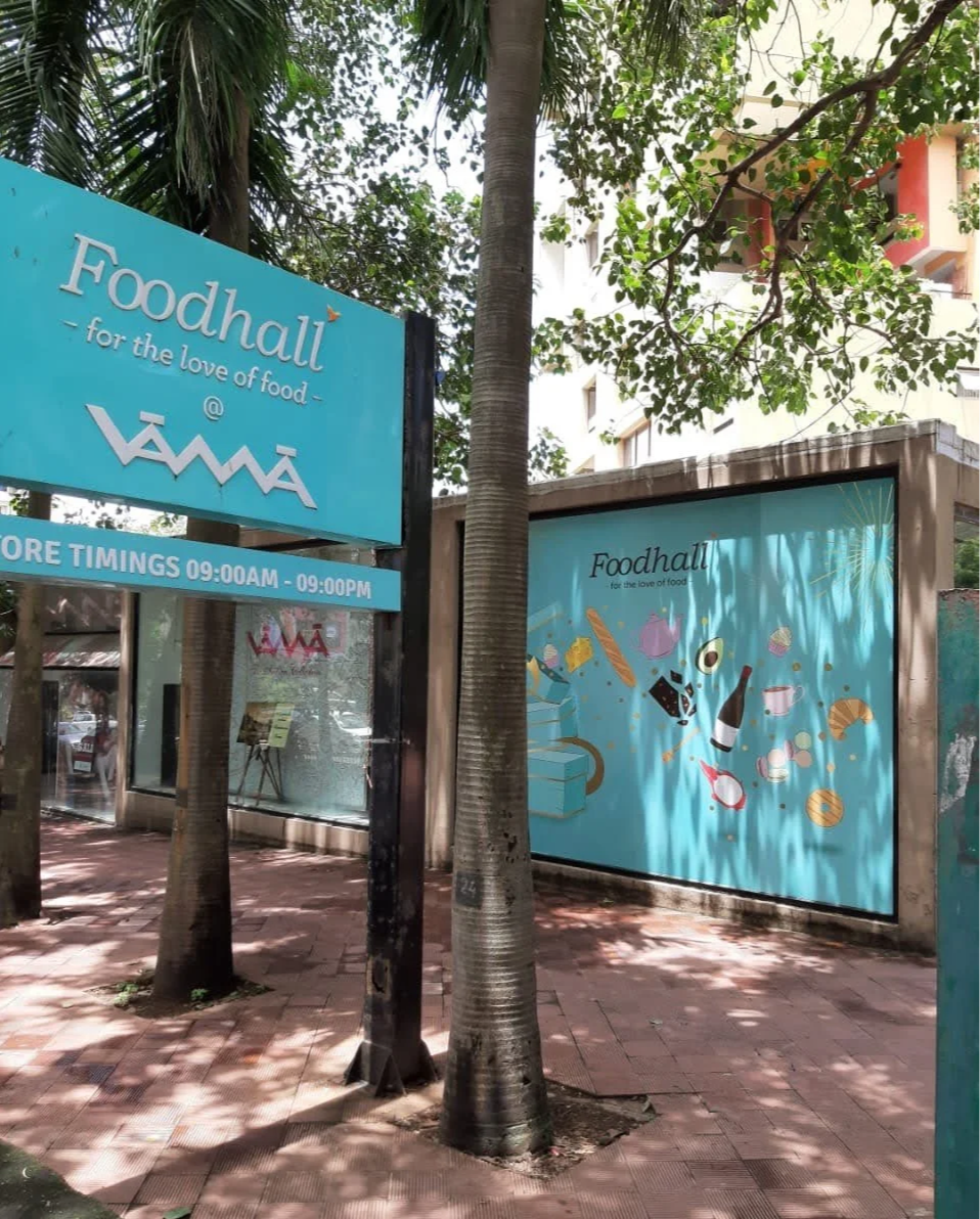 Street view of Foodhall storefront with blue signage and mural, trees, and sidewalk in the daytime.