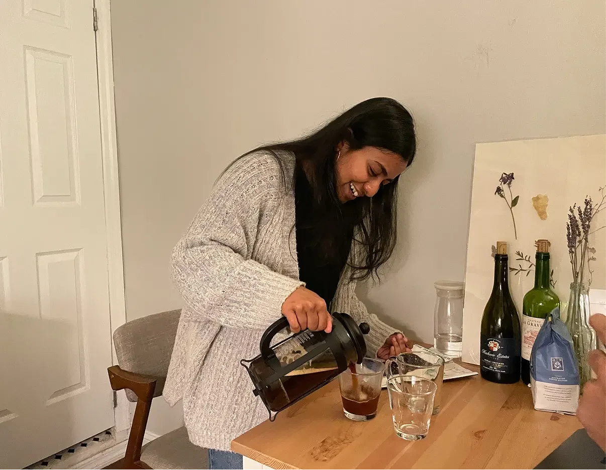 A woman pouring coffee into a glass cup at a dining table with bottles and dried flowers in the background.