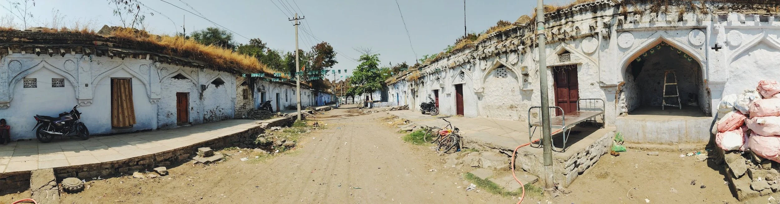 A rural street scene with whitewashed houses, some under construction, in a village. The road is unpaved with construction debris, and there are motorcycles and bicycles parked along the street. Utility poles and wires extend across the scene, and there is a pile of bags on the right side.