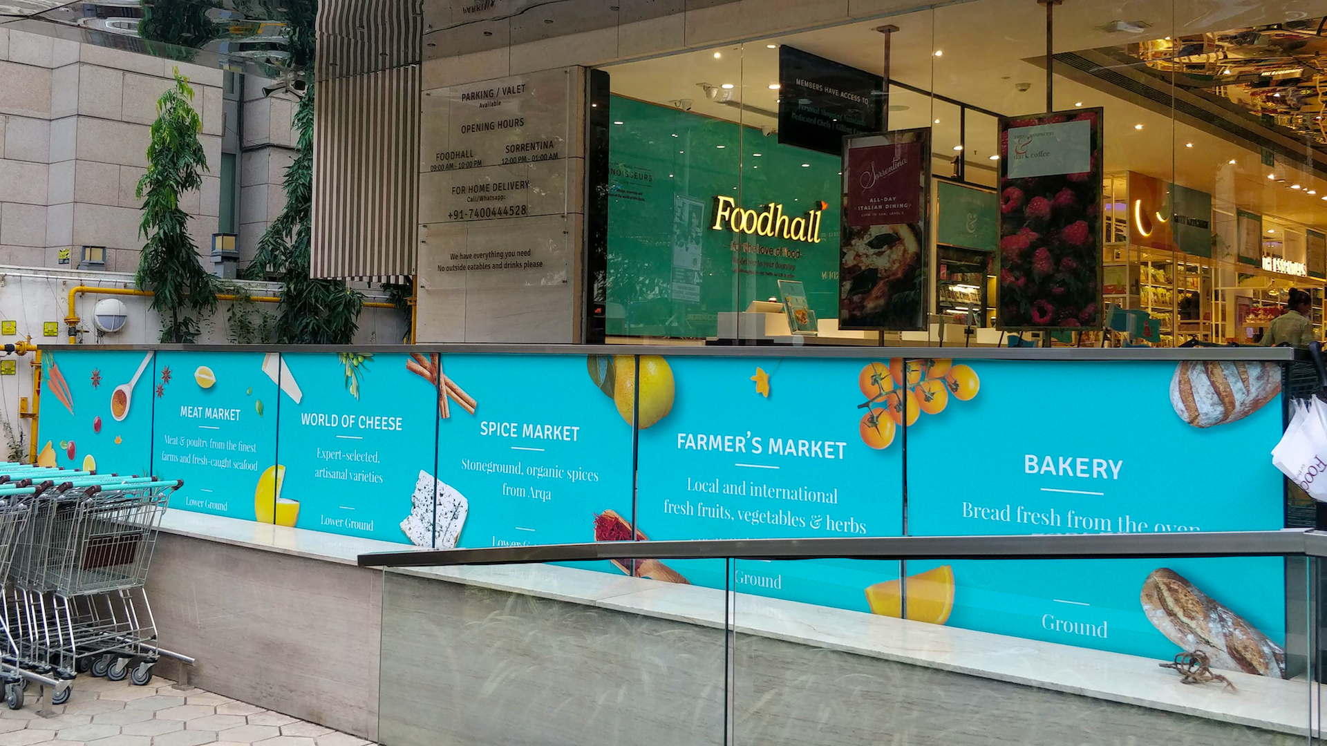 Blue outdoor signboard advertising different markets such as Meat Market, World of Cheese, Spice Market, Farmer’s Market, and Bakery at a shopping mall. The sign features illustrations of vegetables, cheese, spices, fruits, and baked bread.