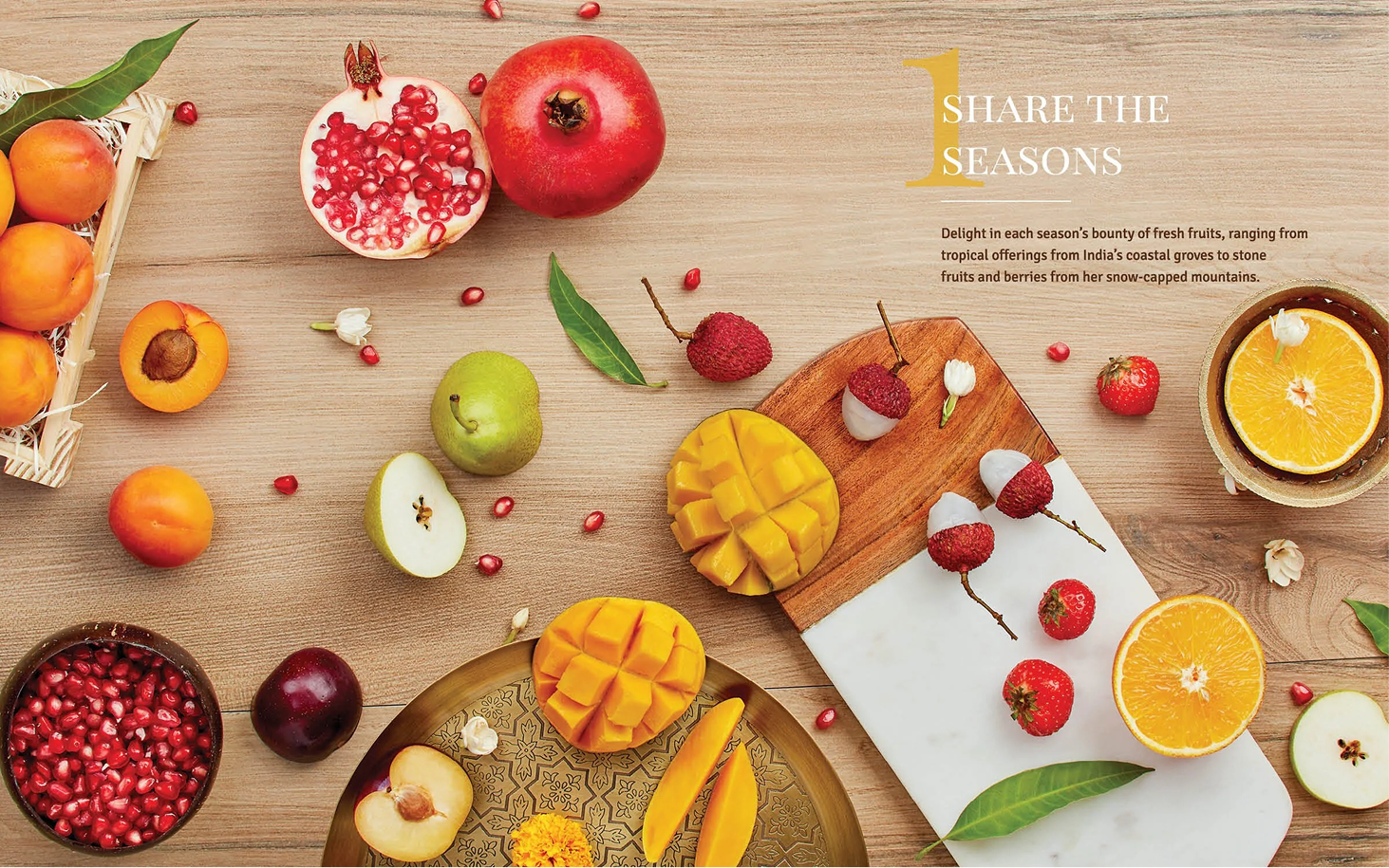 An arrangement of fresh fruits on a wooden surface including pomegranates, apples, strawberries, apricots, a mango, oranges, and lychee, with some cut open and some on plates or cutting boards.