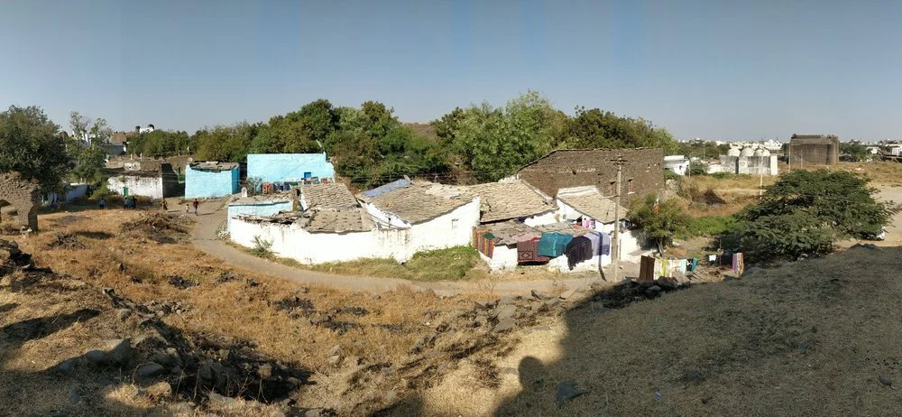 A small village with modest houses, some with clothes hanging outside, surrounded by dry land and sparse trees, under a clear blue sky.