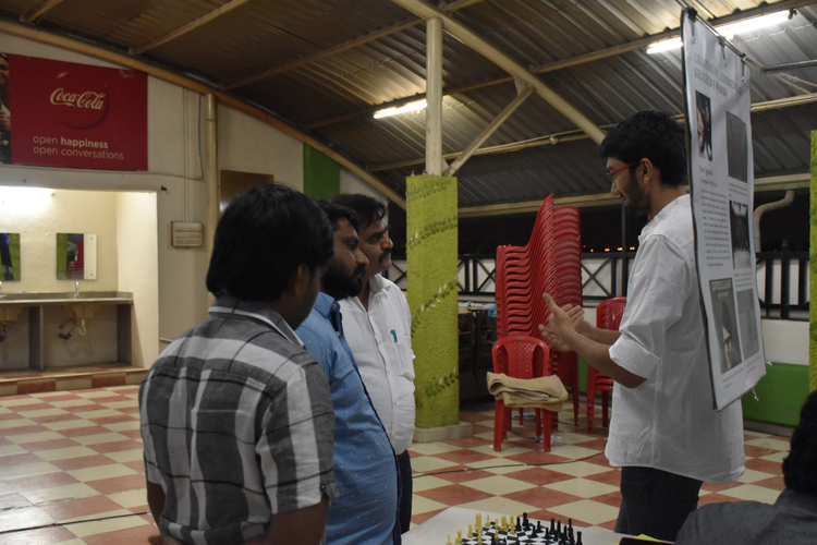 A man is teaching chess to four people in a room with a checkered floor and stacked red chairs.
