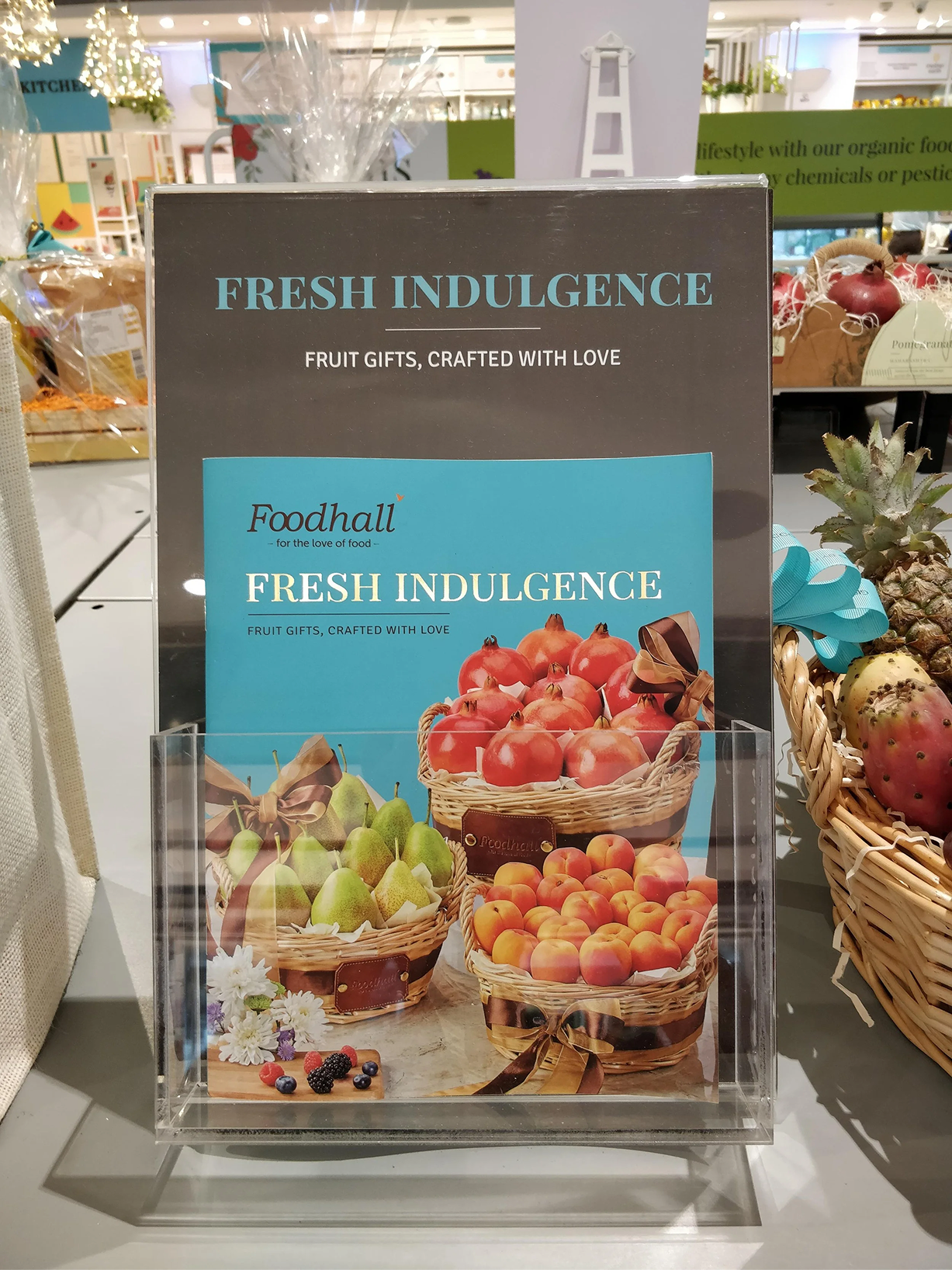 Display sign for Fresh Indulgence fruit gifts at Foodhall, featuring baskets of apples, pears, and peaches arranged on a table, with flowers and berries in front.