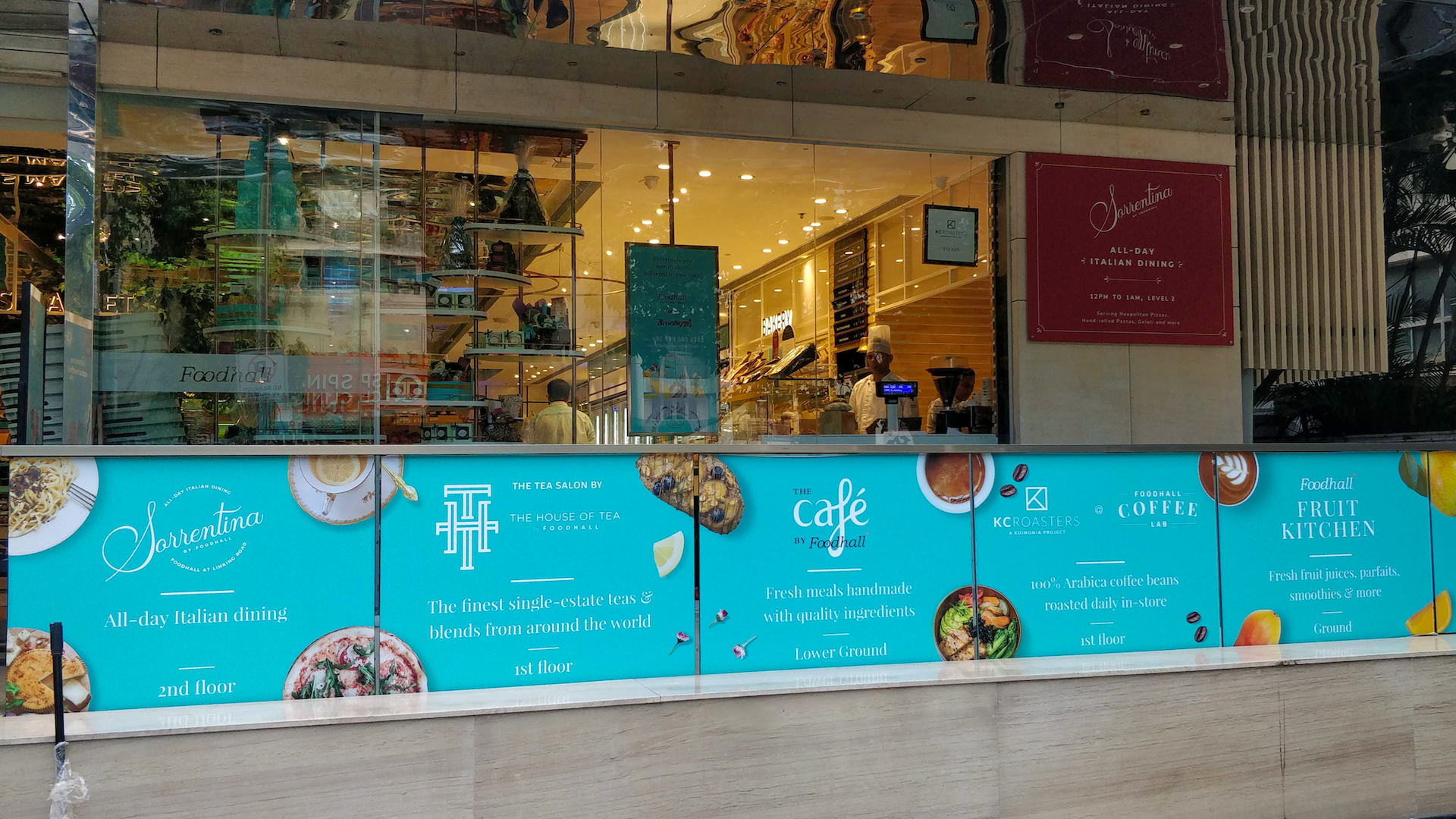 Outside view of a food court with a storefront displaying blue advertisements for Italian dining, tea, cafe, coffee, and fruit kitchen at Foodhall, featuring images of food and drinks.