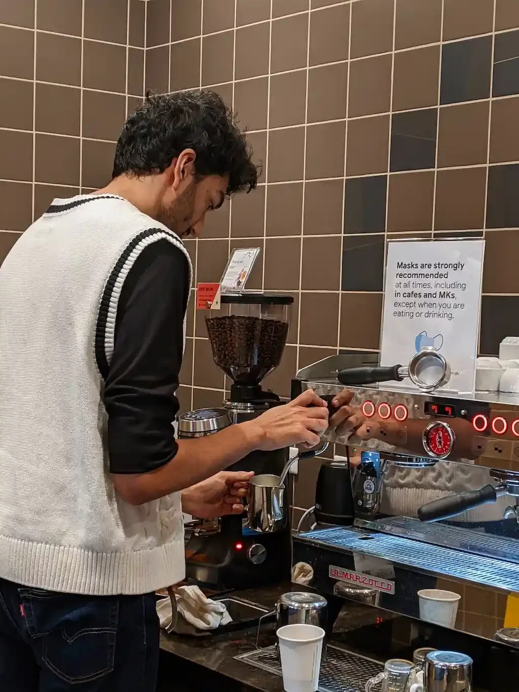 A man making coffee using an espresso machine in a coffee shop. Coffee beans are visible in a grinder behind him, with a sign about mask recommendations on the wall.