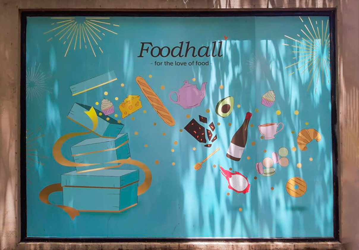 Colorful sign for Foodhall showing various food items like cheese, bread, chocolate, avocado, wine, cupcake, croissant, and macarons, with a blue background and decorative fireworks.