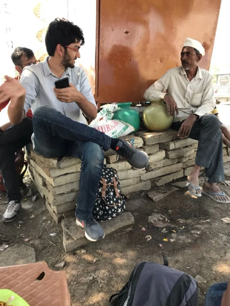Two men sit on a brick and stone bench, one young with dark hair and a beard holding a phone, and one older with gray hair and a cap, next to large jars, in an outdoor setting with a brown door behind them.