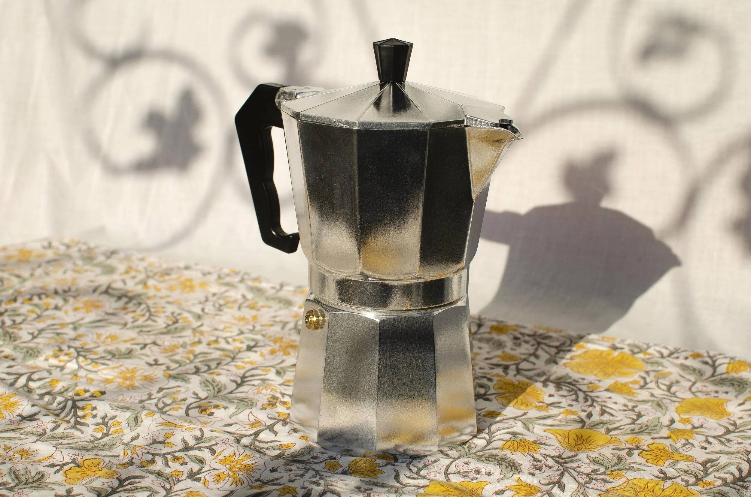 A stovetop moka coffee maker on a floral tablecloth with shadows of leaves cast on a light background.