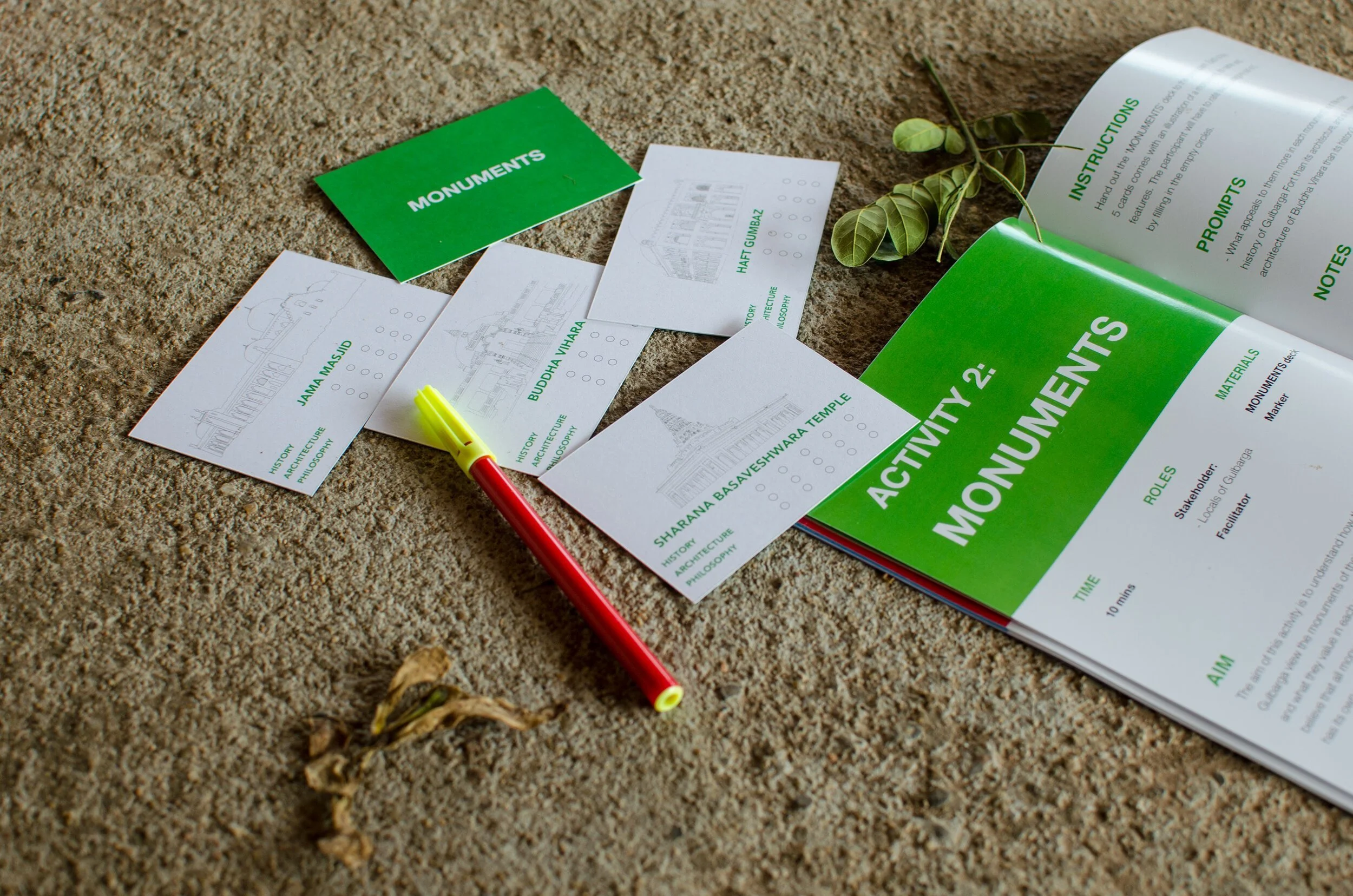 Maps and informational sheets about monuments and activities on a sandy surface, with a red and yellow marker and a small green leafy branch.