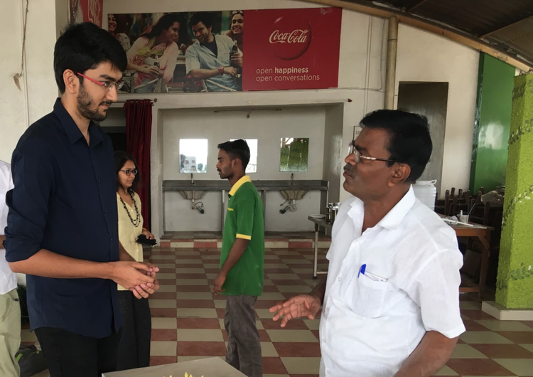 Two men are in conversation in a restaurant or cafe with a checkered floor and a Coca-Cola advertisement on the wall. One man is tall, wearing glasses and a dark shirt, while the other is shorter, wearing glasses and a white shirt. Two other people are in the background, a woman in glasses and a yellow dress, and a man in a green shirt. There are sinks and tables in the background.