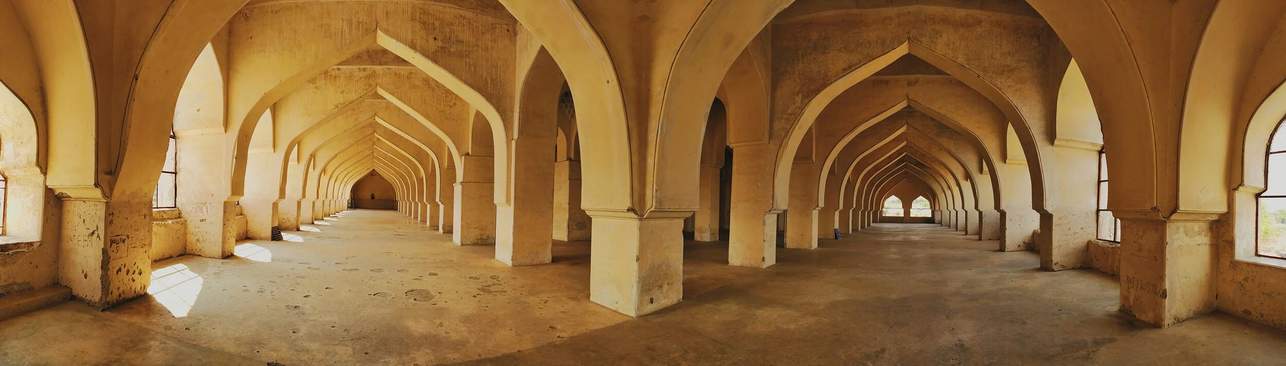 Interior view of an architectural structure with multiple arches, windows lining the sides, and sunlight streaming in through the windows, showing an aged, slightly worn appearance.
