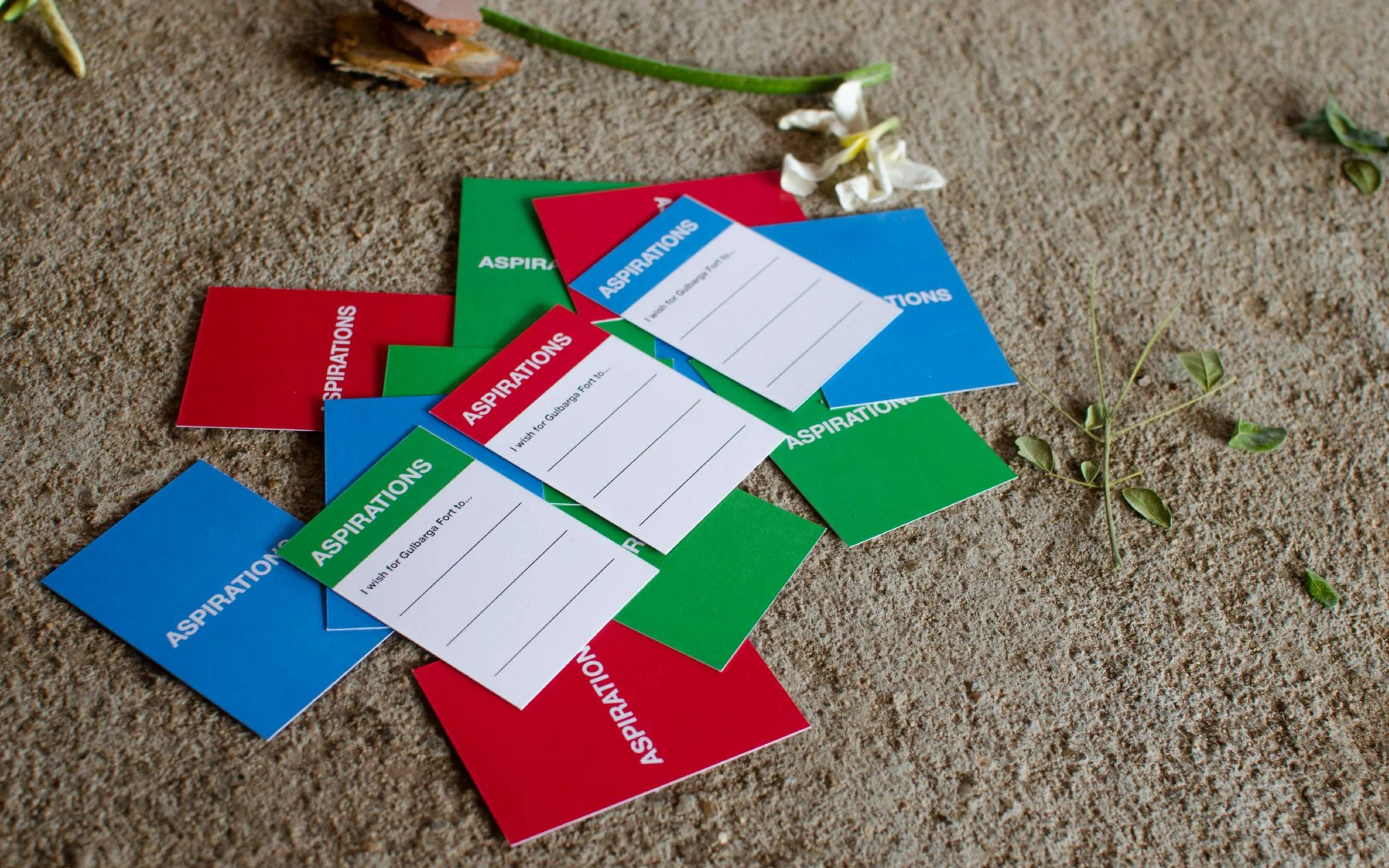 Colorful 'aspirations' and 'dreams' cards scattered on sandy ground with small white flowers and a green plant nearby.