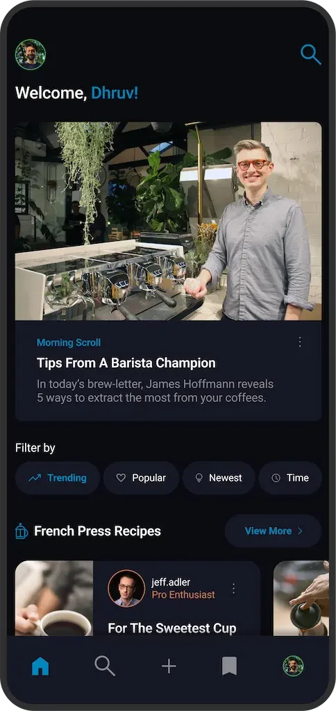 A man with glasses and a gray shirt smiling and standing near a countertop with coffee brewing equipment in a modern cafe or coffee shop.