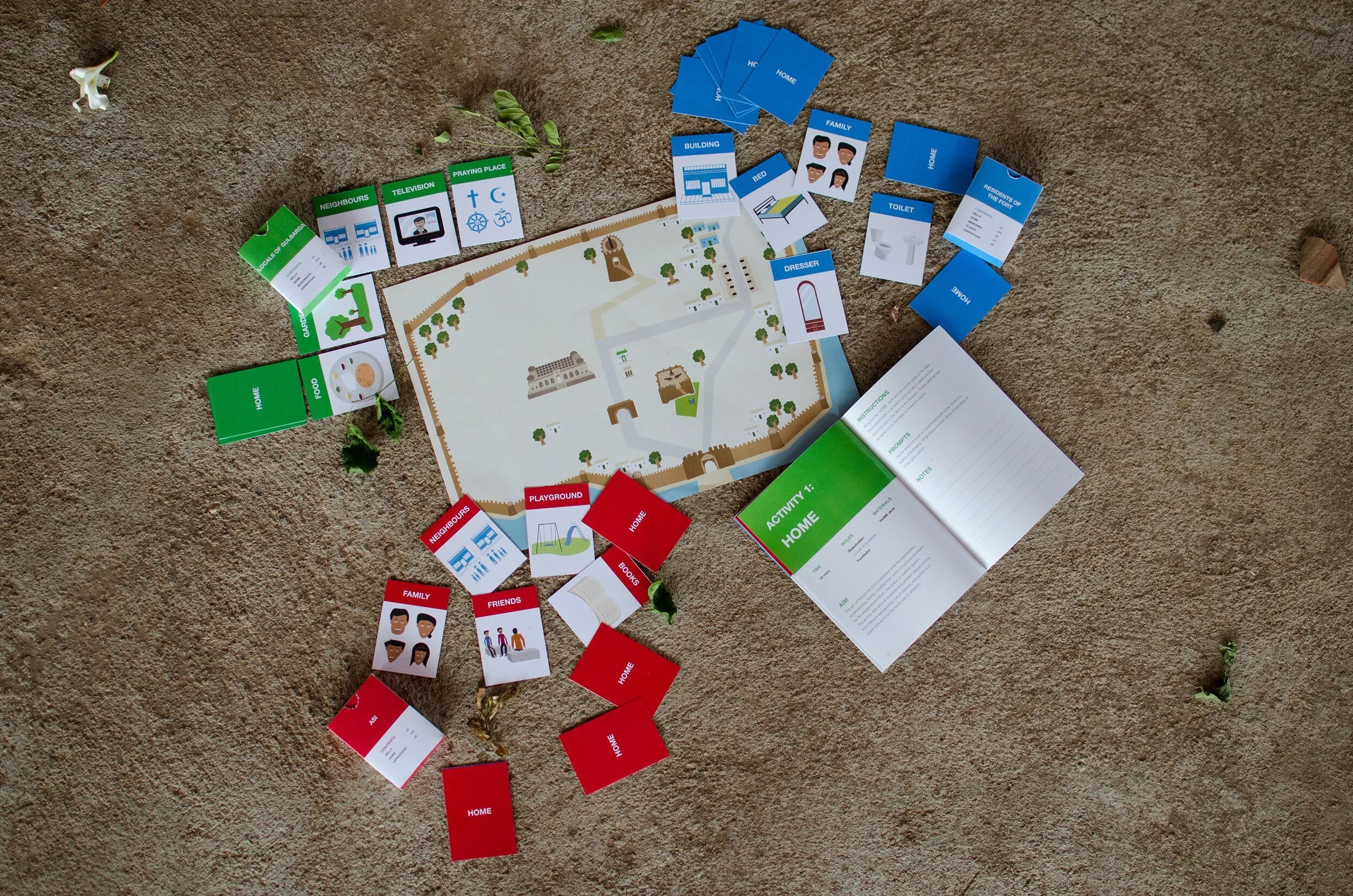 Board game setup on a carpet featuring colorful illustrated tiles and a game book. The tiles represent various home and neighborhood elements, such as family, friends, playground, and specific rooms like kitchen, bedroom, and bathroom, organized around a central map. Some tiles are stacked or scattered around, with leafy plant pieces on the carpet.