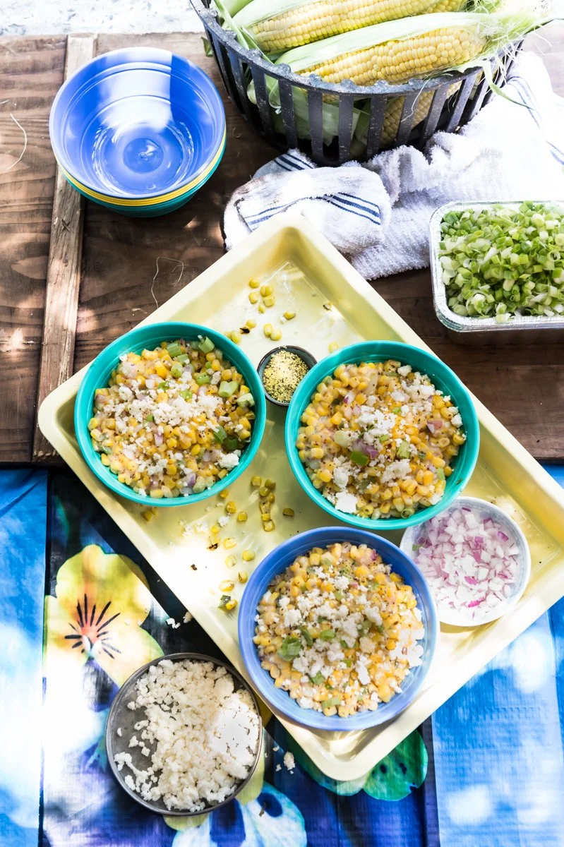 Bowls of Mexican street corn salad on a tray with fresh corn on the cob, chopped green onions, and cheese.