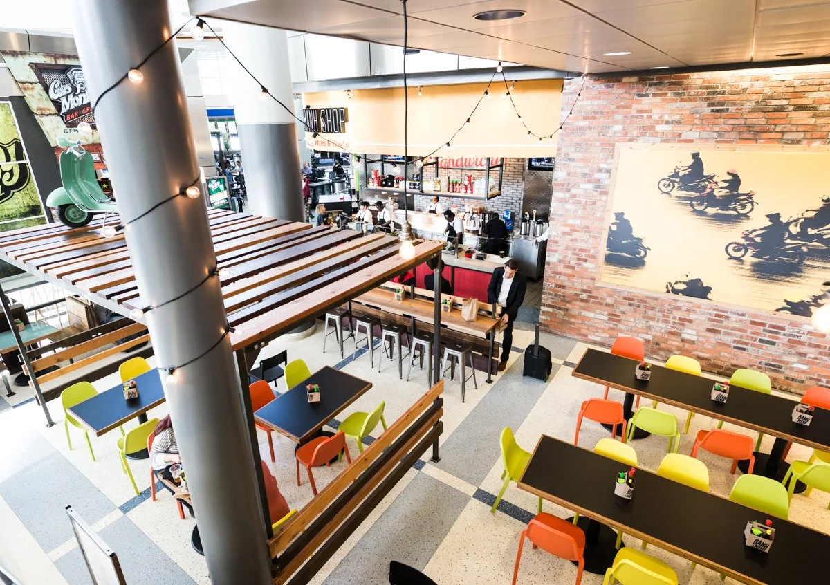 Interior of a modern cafe with colorful chairs, wooden tables, an overhead wooden structure with string lights, a scooter decoration, and a brick wall featuring a motorcycle mural. People are seated and standing around the counter area.