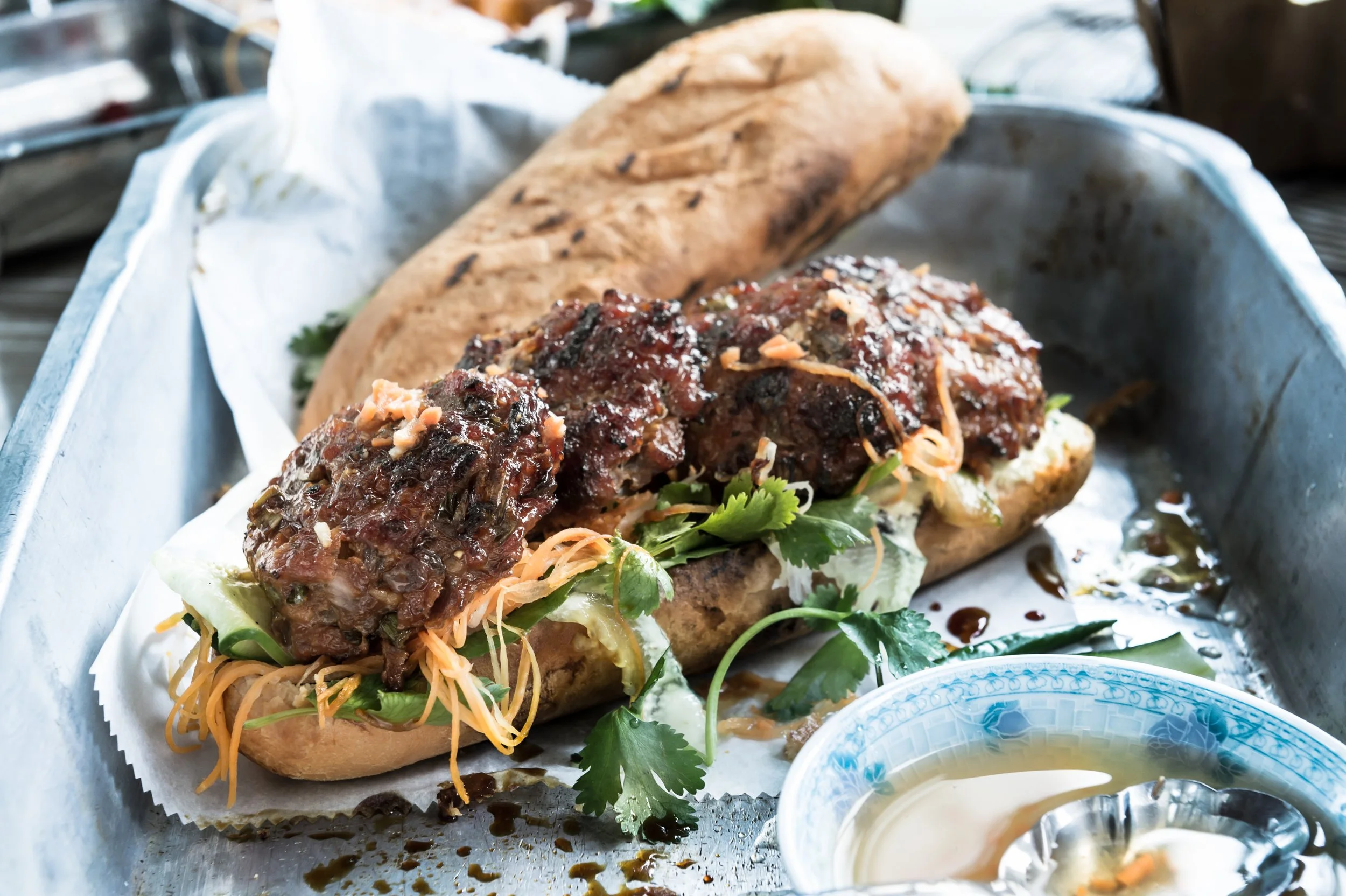 Banh mi sandwich with grilled meat, herbs, and pickled vegetables on a tray with dipping sauce.